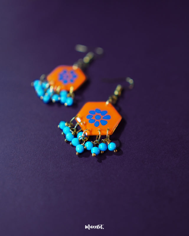 Surkhshaam orange floral stud drop earrings, set against a deep purple background, featuring vibrant blue floral accents and a graceful drop design that beautifully combine boldness and elegance — Muselot.