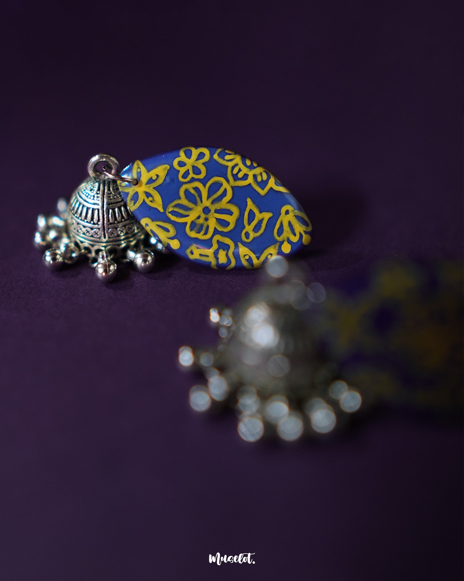 Tabeer blue jhumkas rest gracefully against a subtle purple background, highlighting the floral patterns and intricate detailing that add a timeless elegance to the design — Muselot.