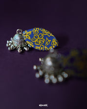 Tabeer blue jhumkas rest gracefully against a subtle purple background, highlighting the floral patterns and intricate detailing that add a timeless elegance to the design — Muselot.