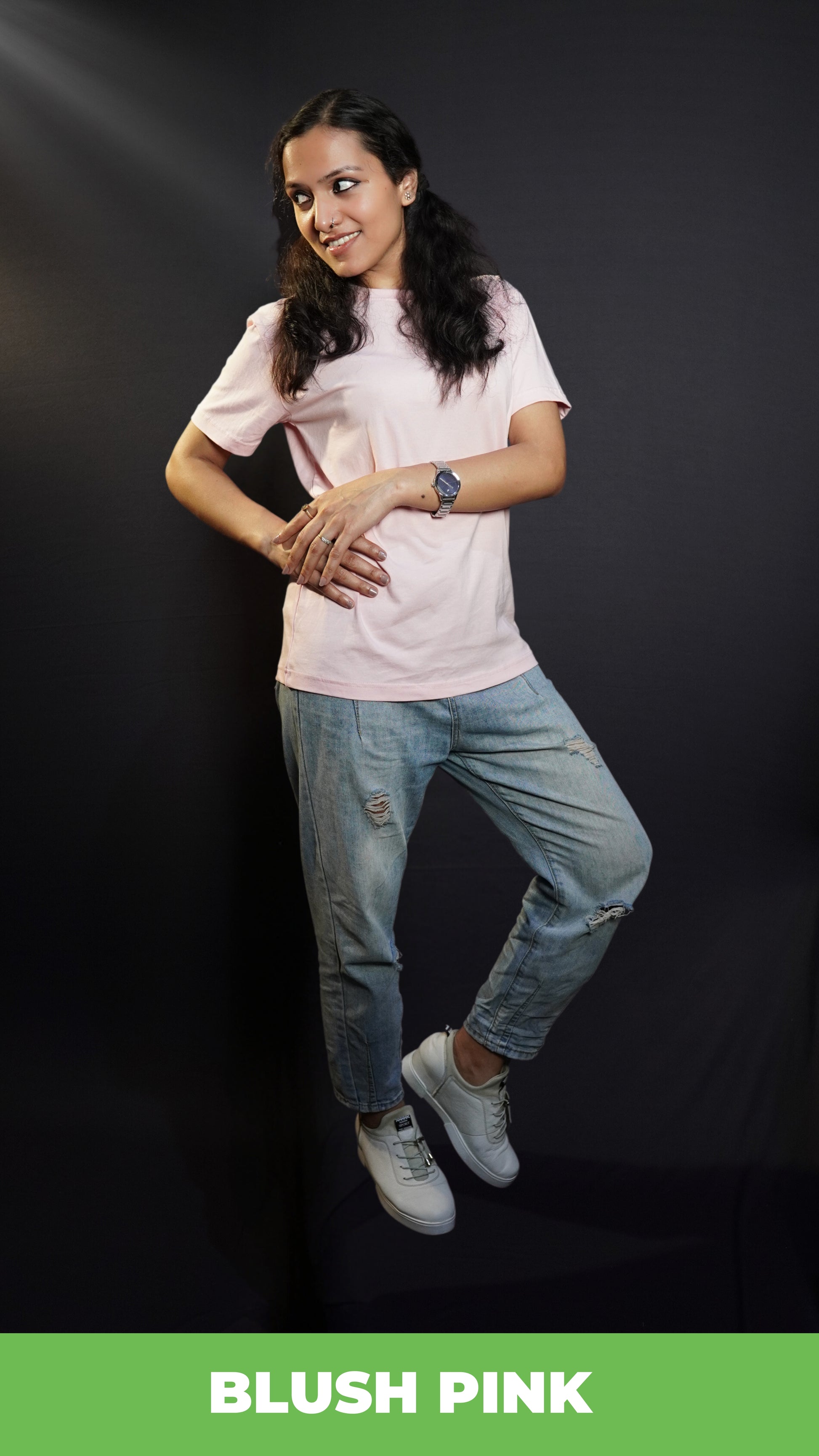 A model poses casually in a blush pink plain t-shirt, blue ripped jeans, and white shoes, placing one hand on her waist and the other on her hand, showcasing a fashionable trendy look-muselot