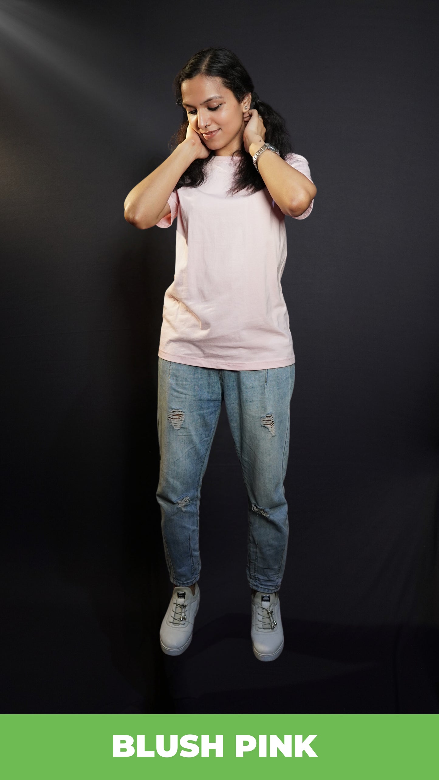 Model in a blush pink t-shirt and light blue ripped jeans, posing with hands on her neck, against a dark background showcasing the casual and trendy fashion styles-muselot