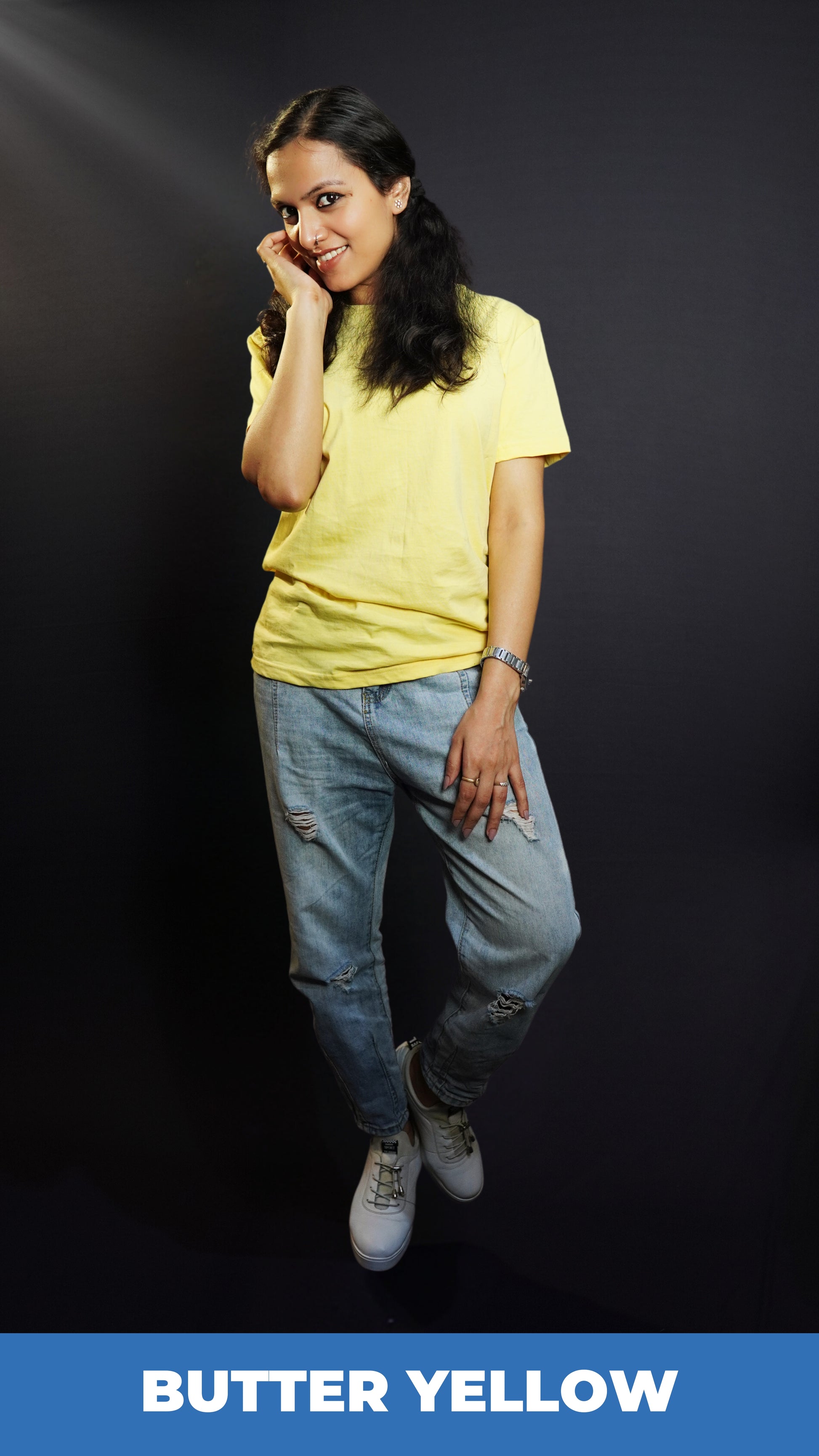 A woman posing happily as she stands against a dark background with her hand on the side of her face and another on her thigh, wearing a butter yellow plain long length stretchable t-shirt, showcasing a voguish popular style-Muselot