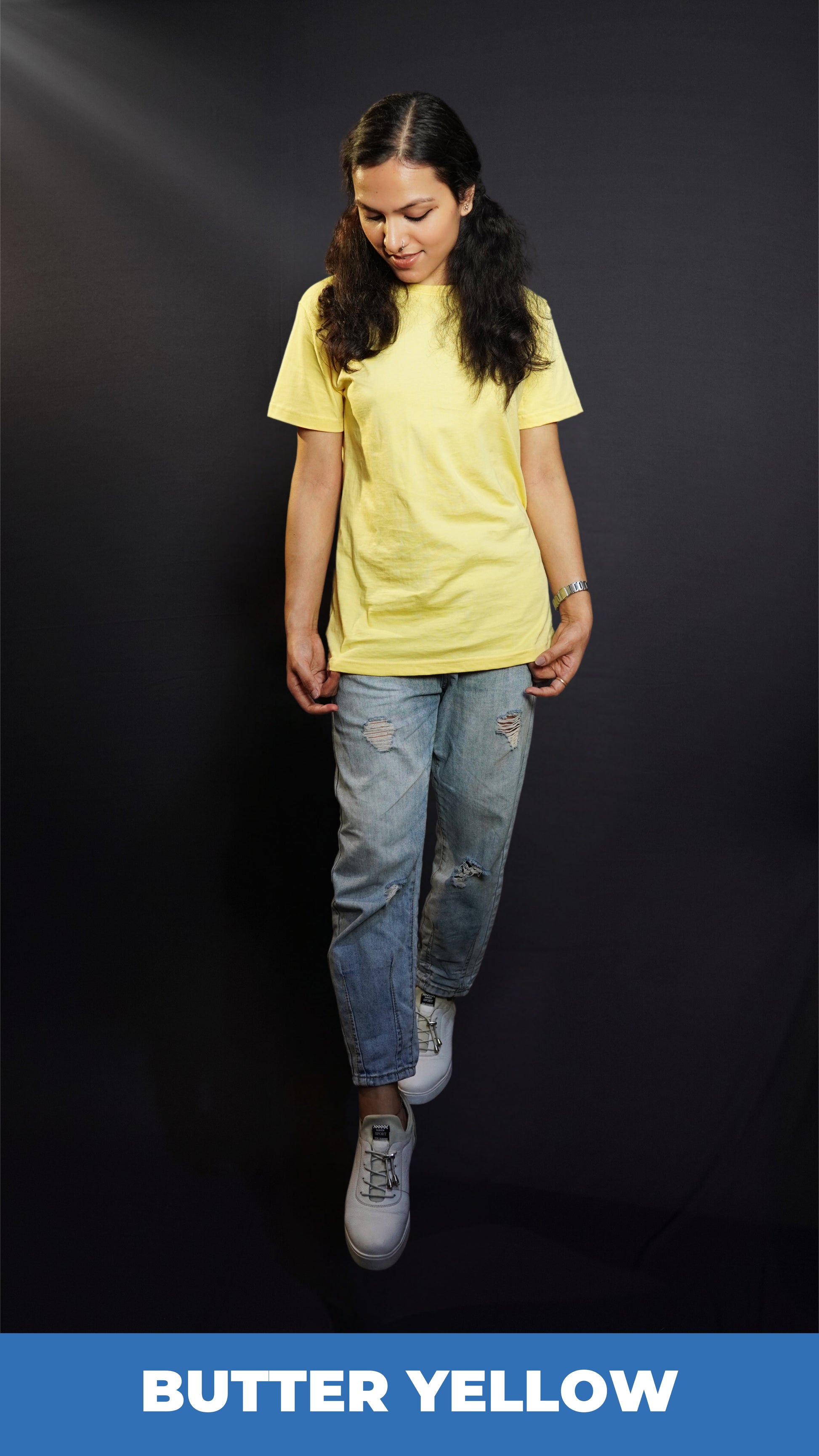 a stylish model wearing a butter yellow plain stretchable t-shirt, standing with her head slightly bent down and her hands grabbing the side of the t-shirt, showcasing an elegant casual fit-Muselot