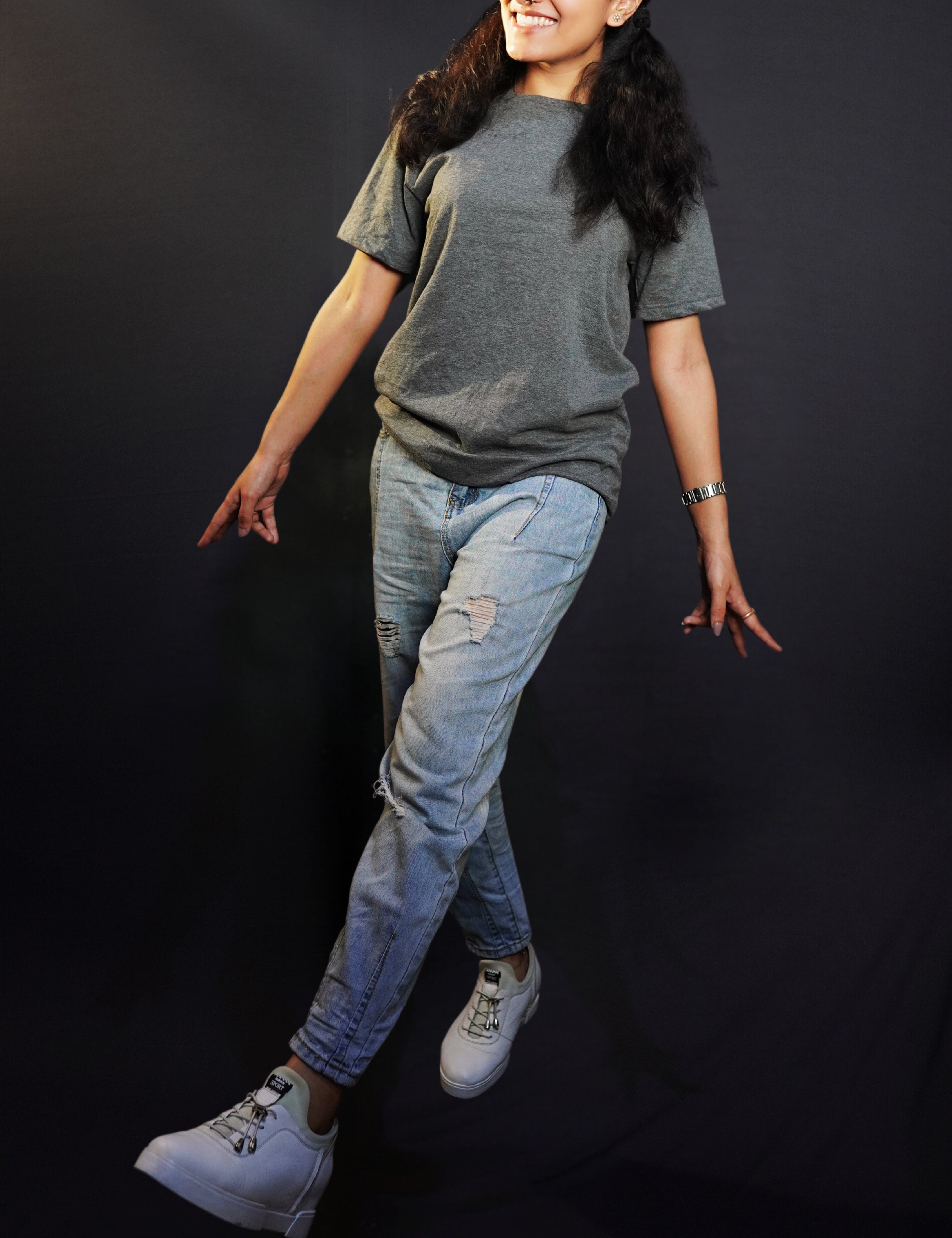 a happy woman walking wearing a charcoal grey t shirt with ripped jeans and white sports shoes with a smile on her face against a dark background-muselot