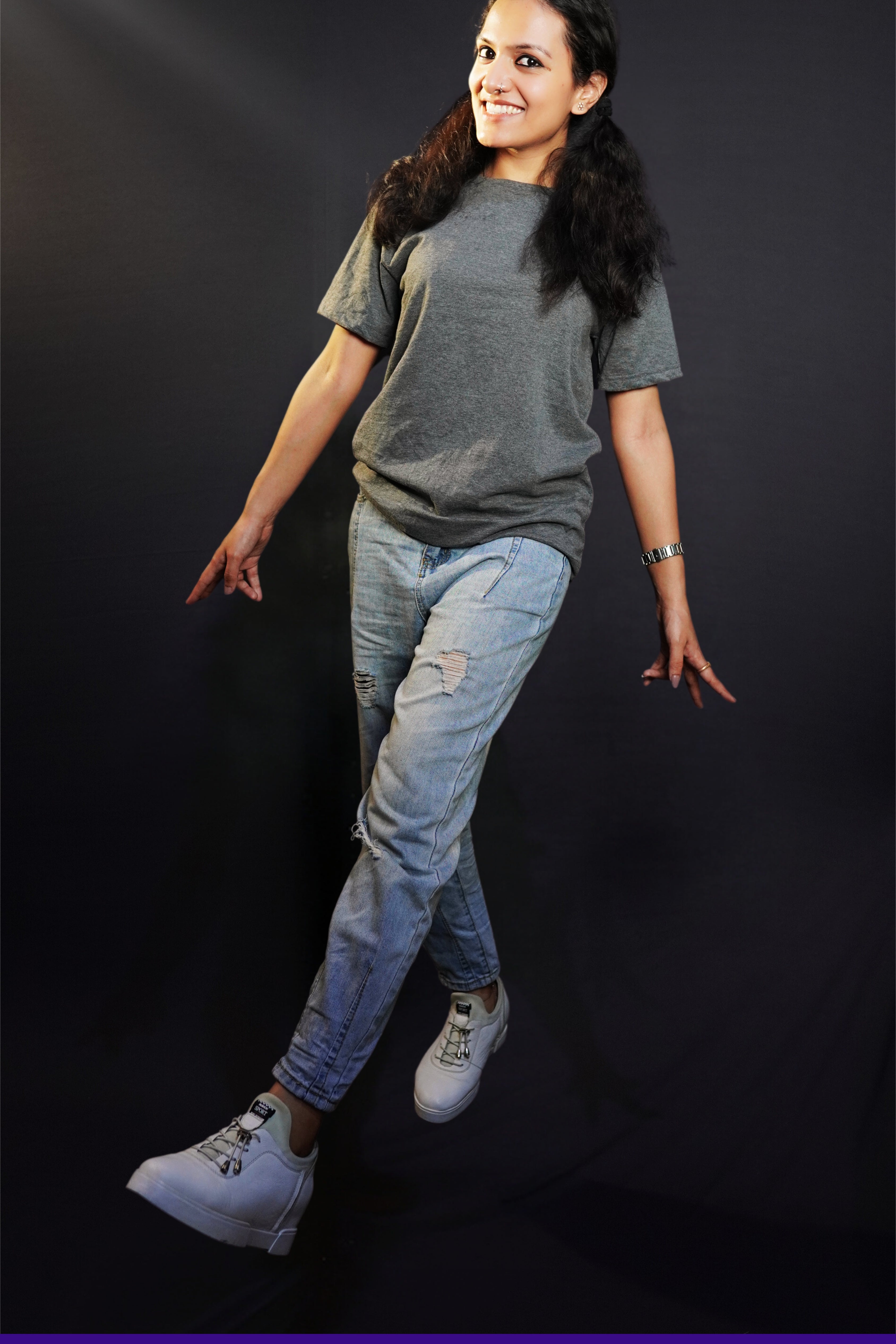 a happy woman walking wearing a charcoal grey t shirt with ripped jeans and white sports shoes with a smile on her face against a dark background-muselot