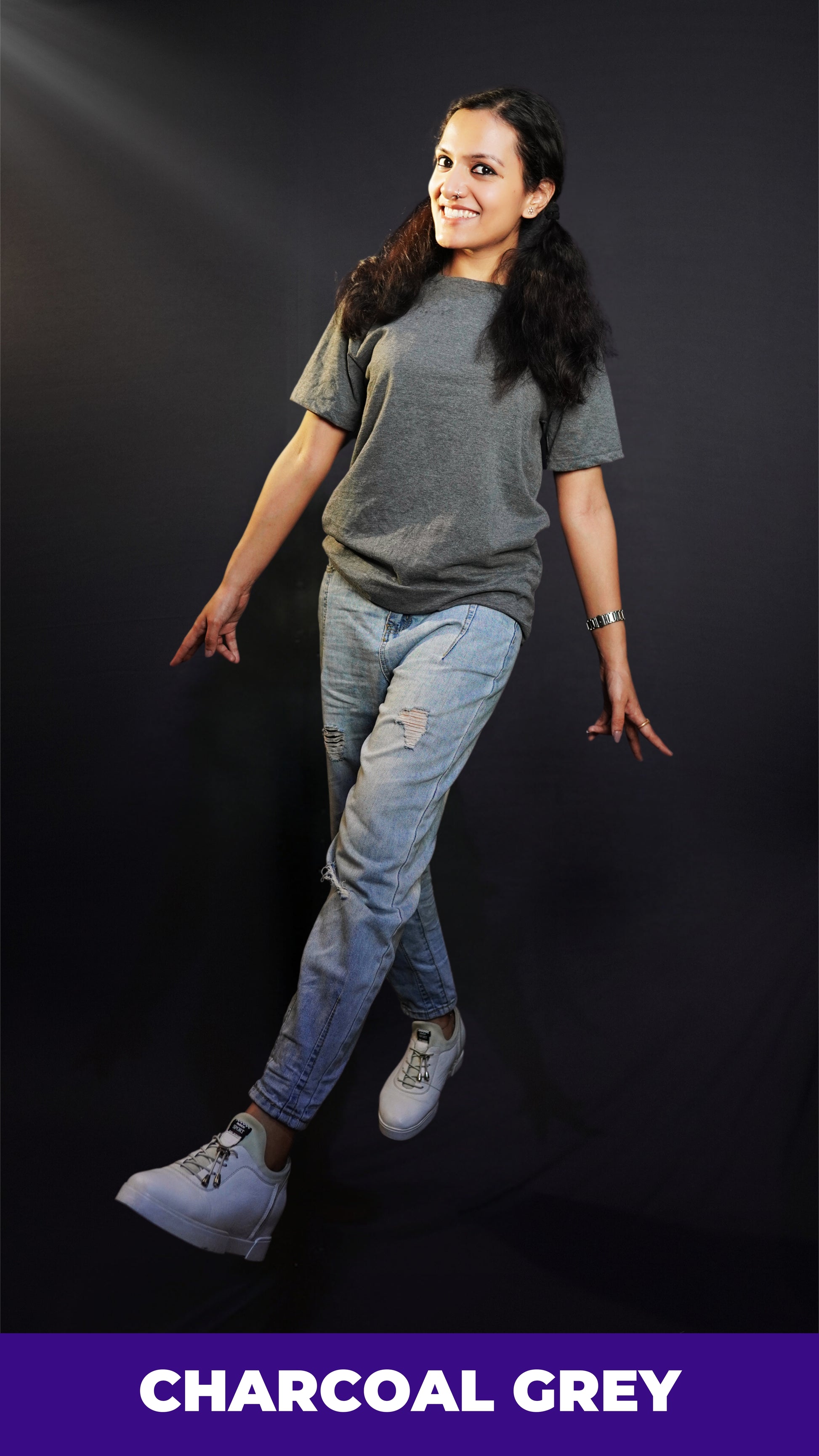 a happy woman walking wearing a charcoal grey t shirt with ripped jeans and white sports shoes with a smile on her face against a dark background-muselot