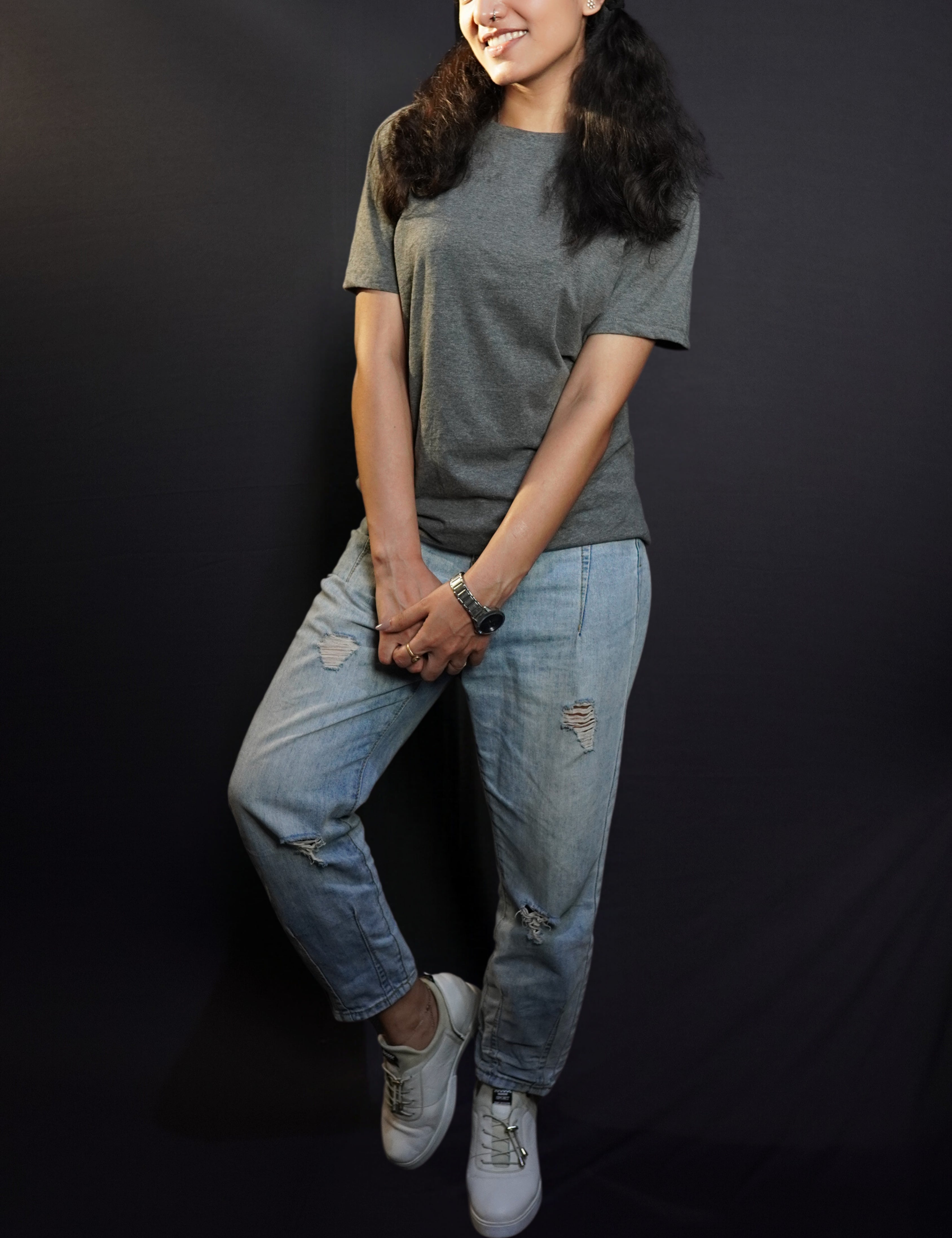 a woman with a smile on her face wearing long length t shirt pairing with ripped jeans and white shoes and a silver watch around her wrist-muselot