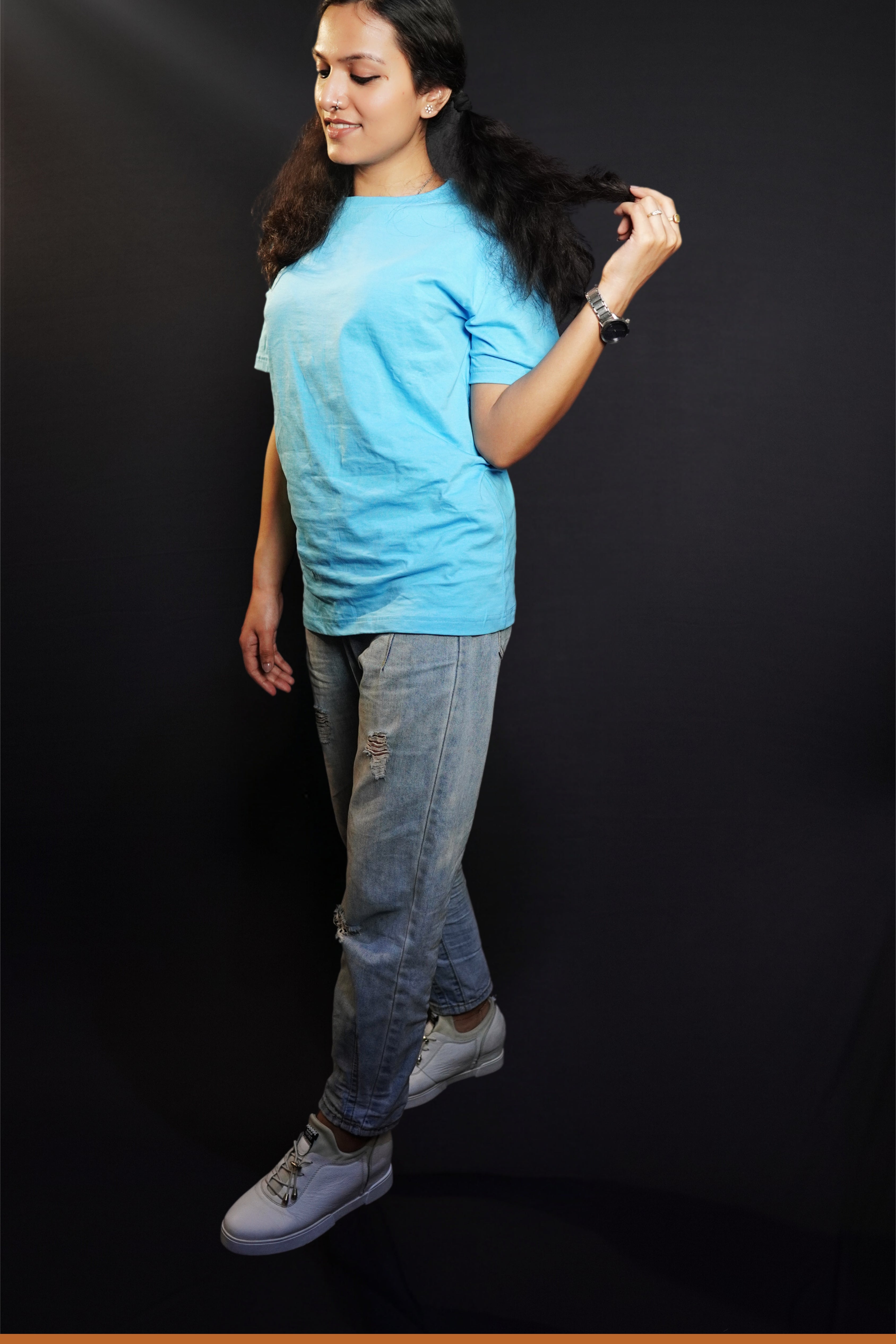 A stylish model standing sideways, pulling a tip of her hair, wearing a plain cloudless blue round neck t shirt showcasing a casual comfortable look-Muselot