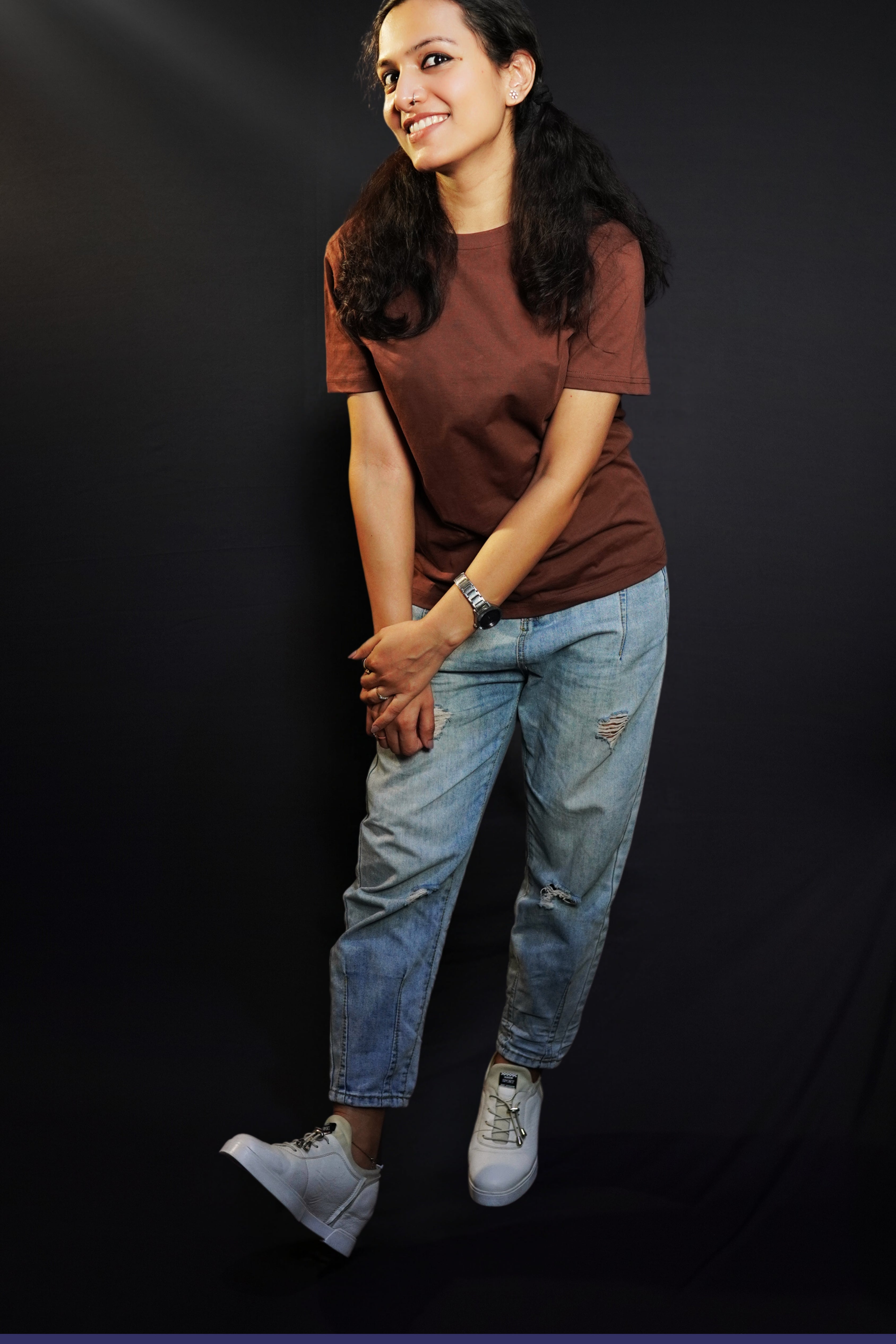 A woman in a long length plain coffee brown t-shirt, posing happily as she slightly bends forward with her hands grabbing each other, highlighting a popular cozy fit-Muselot