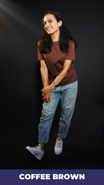 A woman in a long length plain coffee brown t-shirt, posing happily as she slightly bends forward with her hands grabbing each other, highlighting a popular cozy fit-Muselot