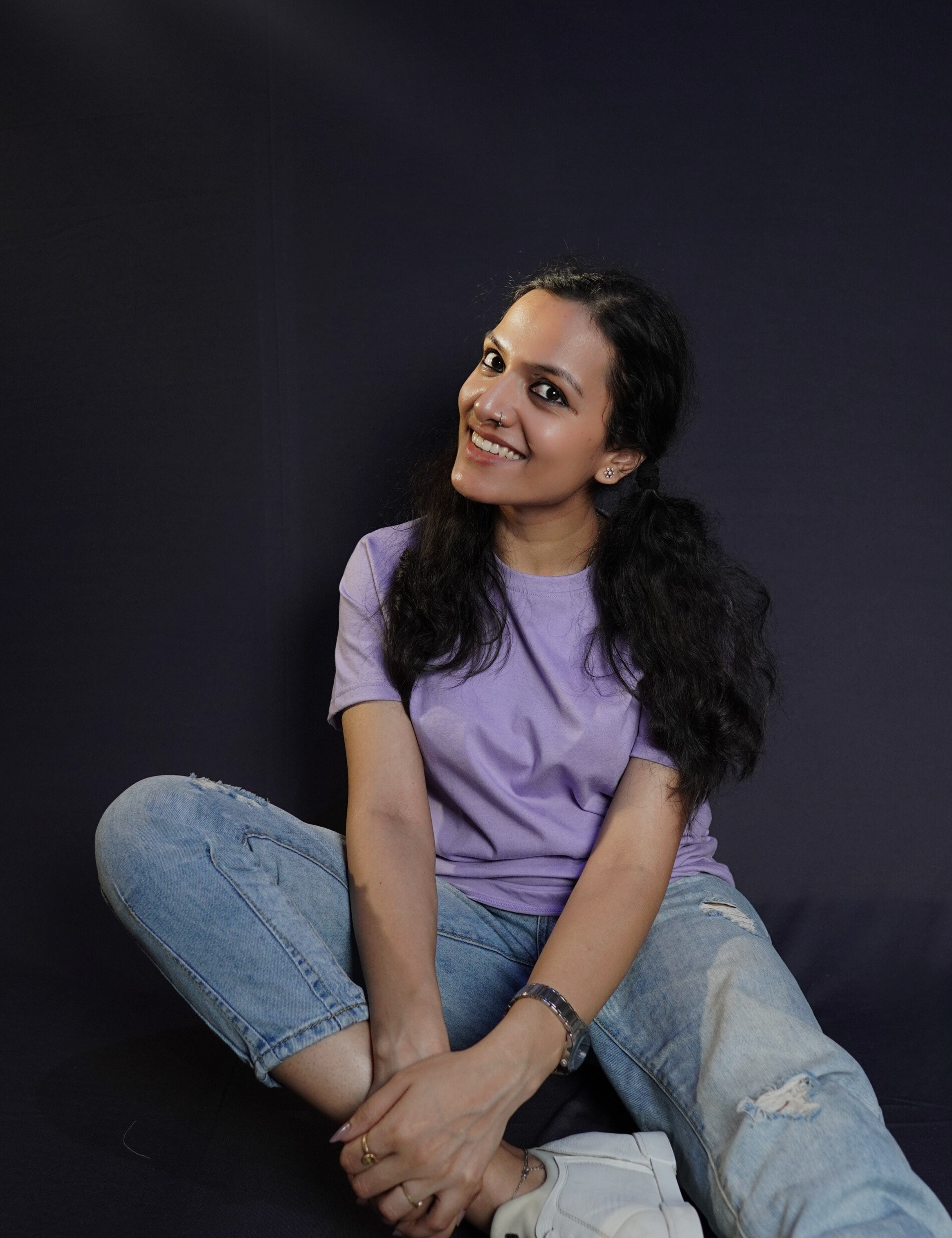 A stylish model wearing a plain short sleeved round neck t-shirt, posing by sitting against a dark background grabbing her leg gently with both hands, showcasing a swank perfect fit-Muselot