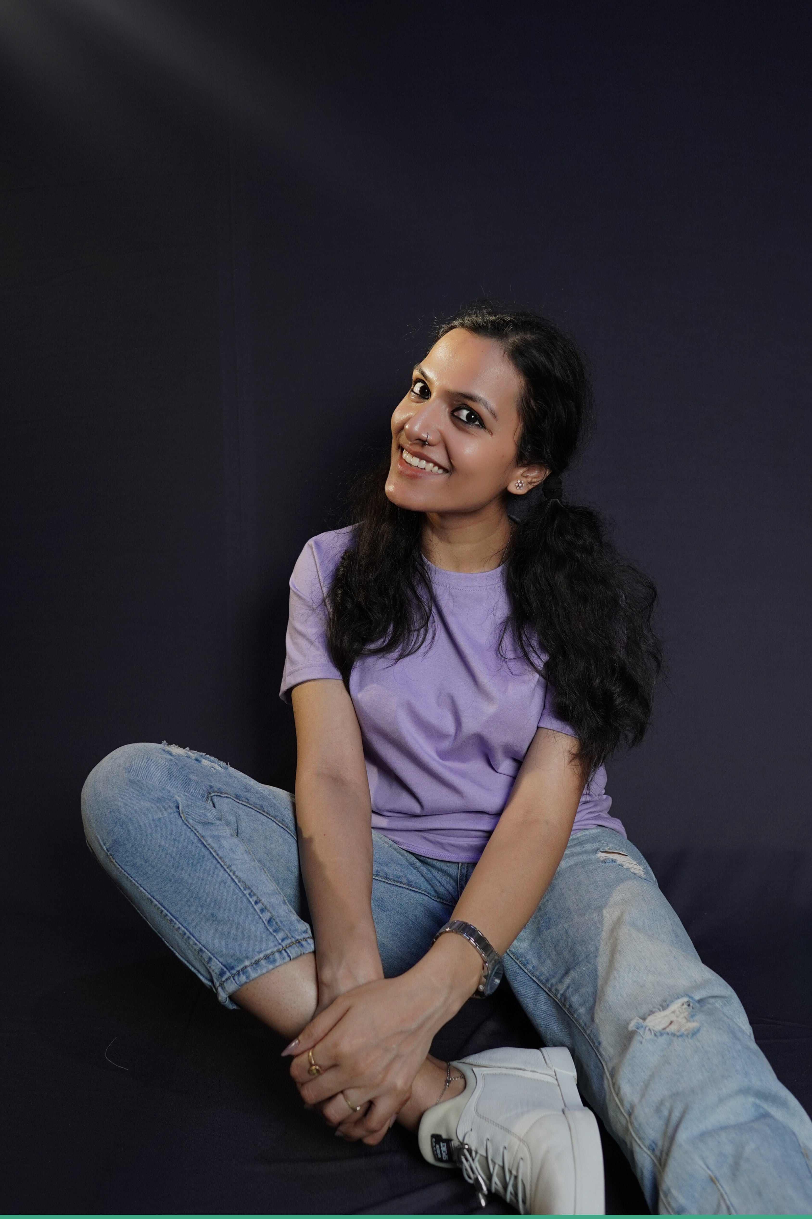 A stylish model wearing a plain short sleeved round neck t-shirt, posing by sitting against a dark background grabbing her leg gently with both hands, showcasing a swank perfect fit-Muselot