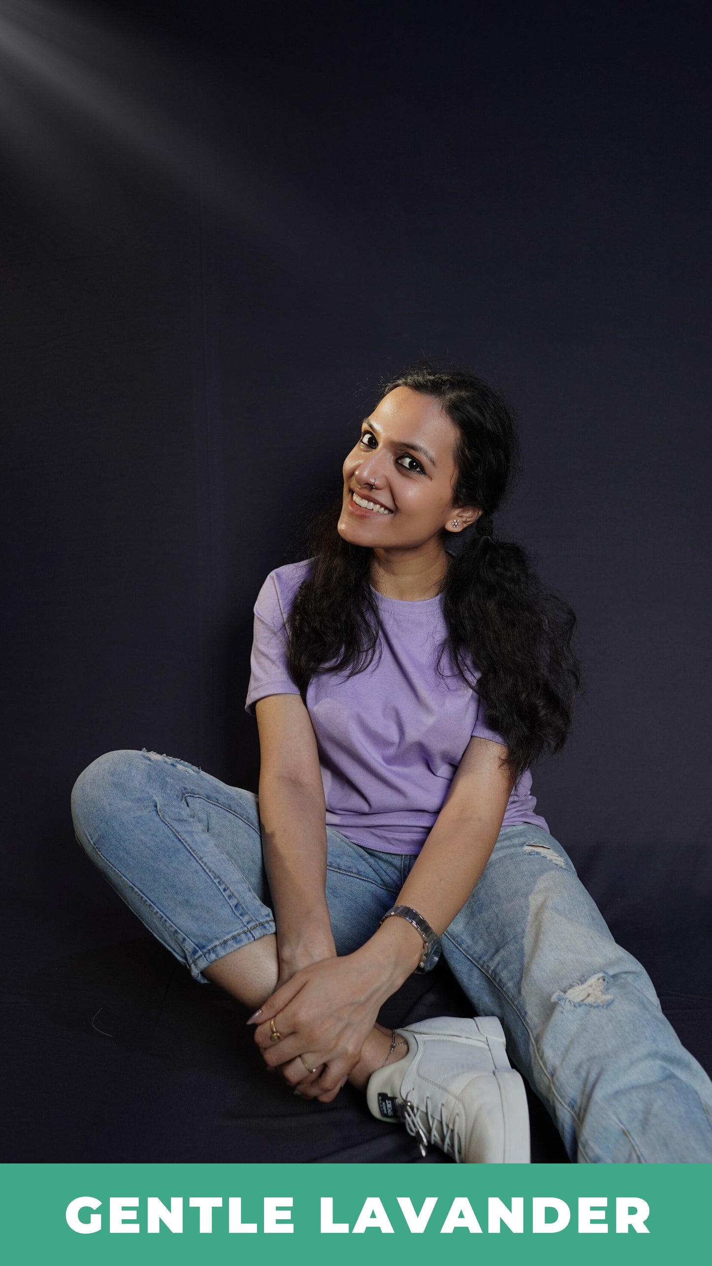 A stylish model wearing a plain short sleeved round neck t-shirt, posing by sitting against a dark background grabbing her leg gently with both hands, showcasing a swank perfect fit-Muselot