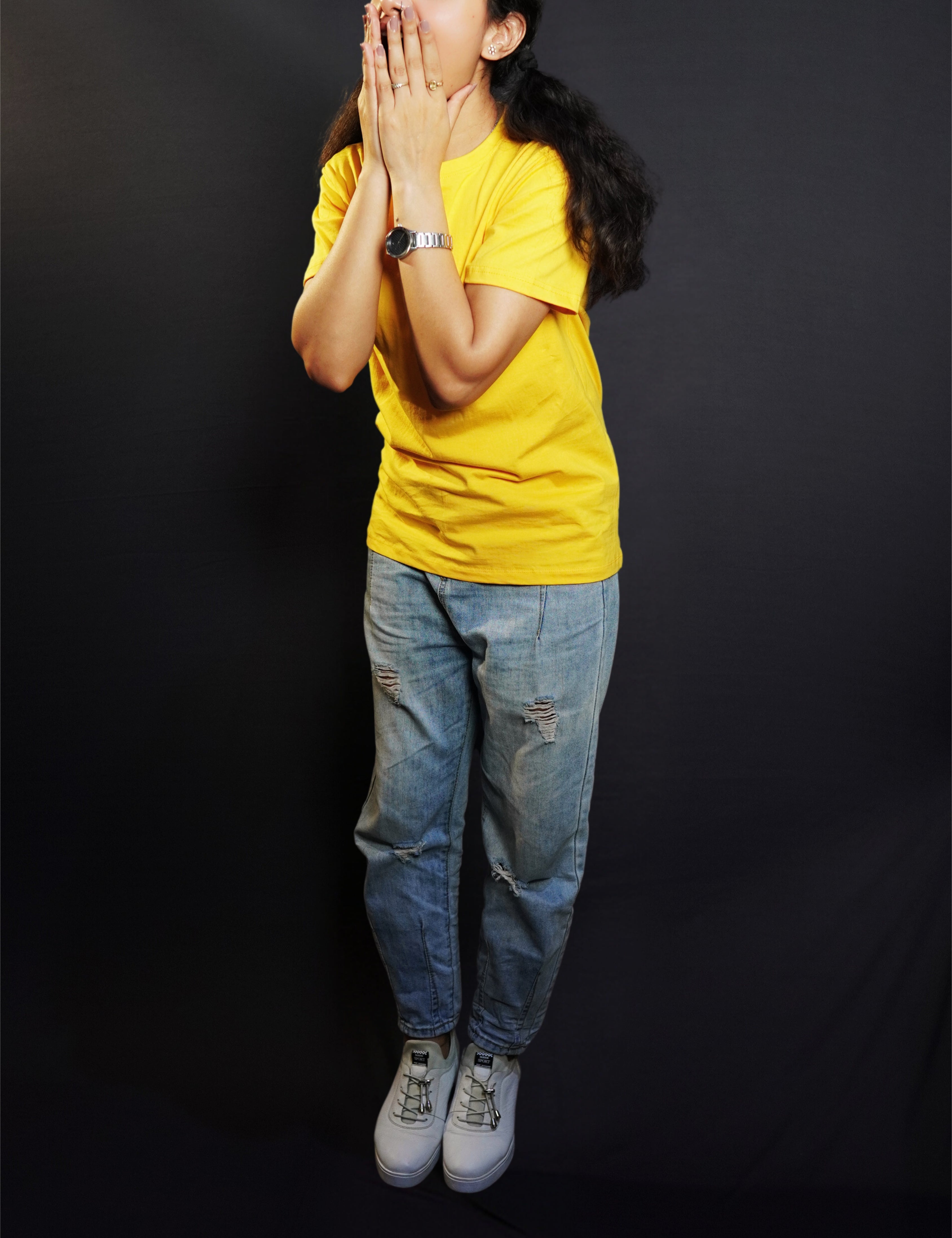 A woman wearing a unisex golden yellow long length T shirt pairing with a blue jeans and sports shoes standing excitedly against a dark background-Muselot