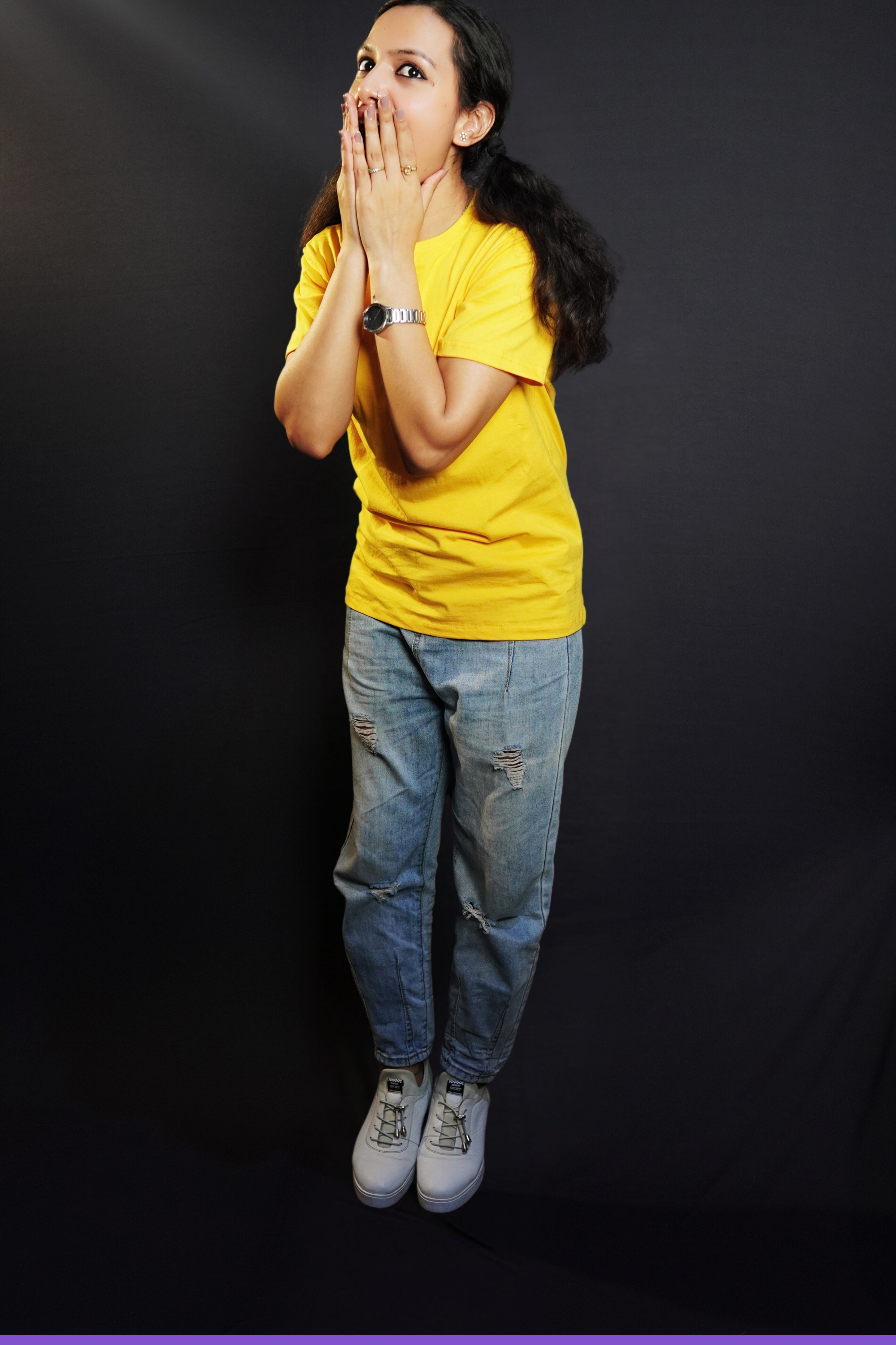 A woman wearing a unisex golden yellow long length T shirt pairing with a blue jeans and sports shoes standing excitedly against a dark background-Muselot