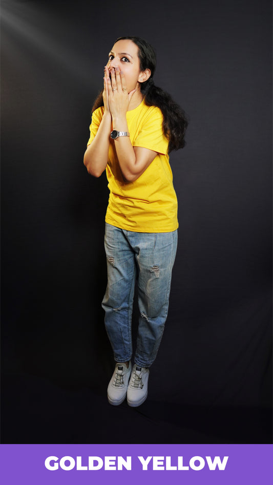 A woman wearing a unisex golden yellow long length T shirt pairing with a blue jeans and sports shoes standing excitedly against a dark background-Muselot