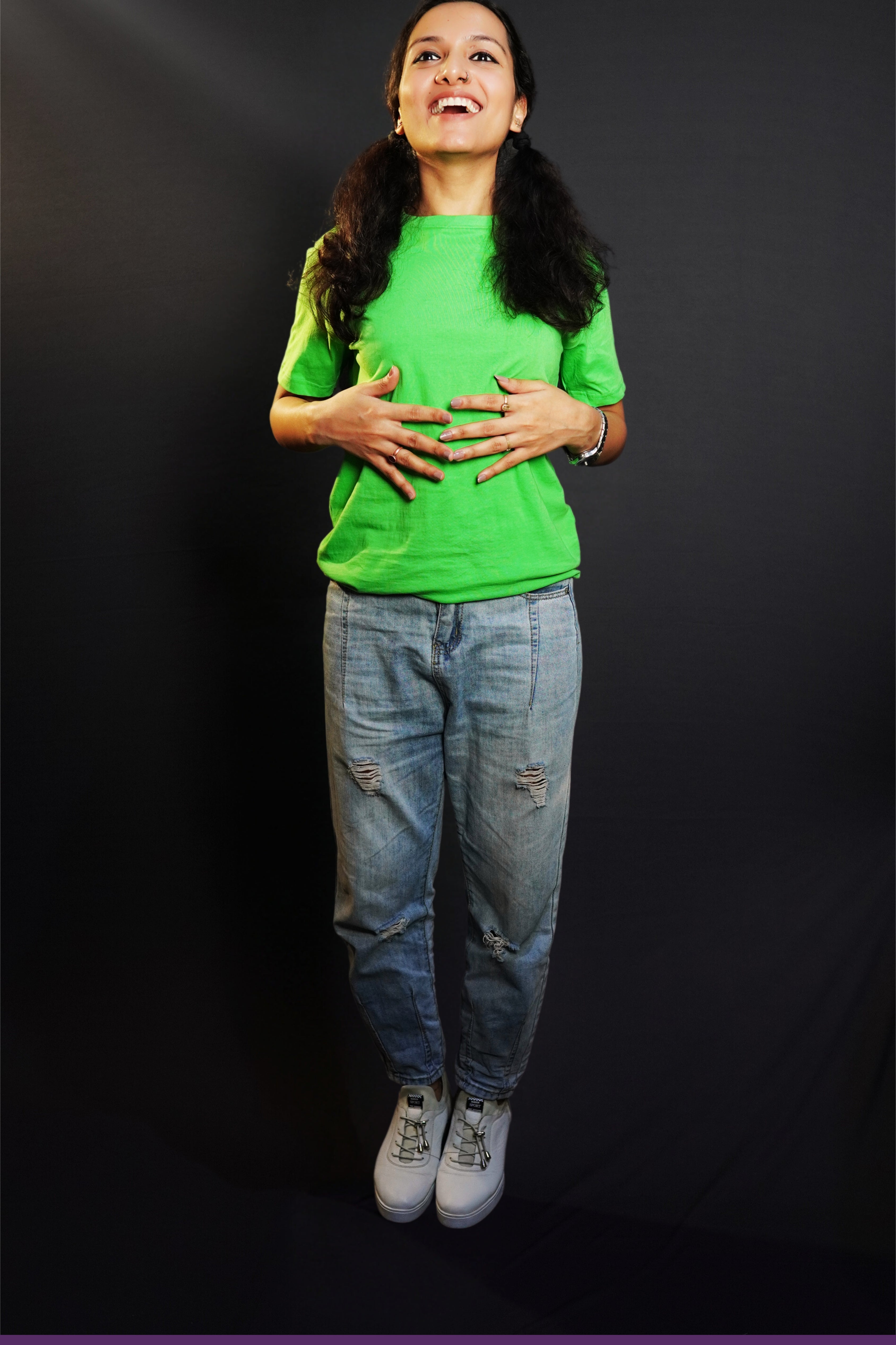 A woman laughing happily standing against a dark background as her hands gently lie on her stomach, wearing a liril green plain unisex round neck short sleeves t-shirt, showcasing a casual everyday look-Muselot