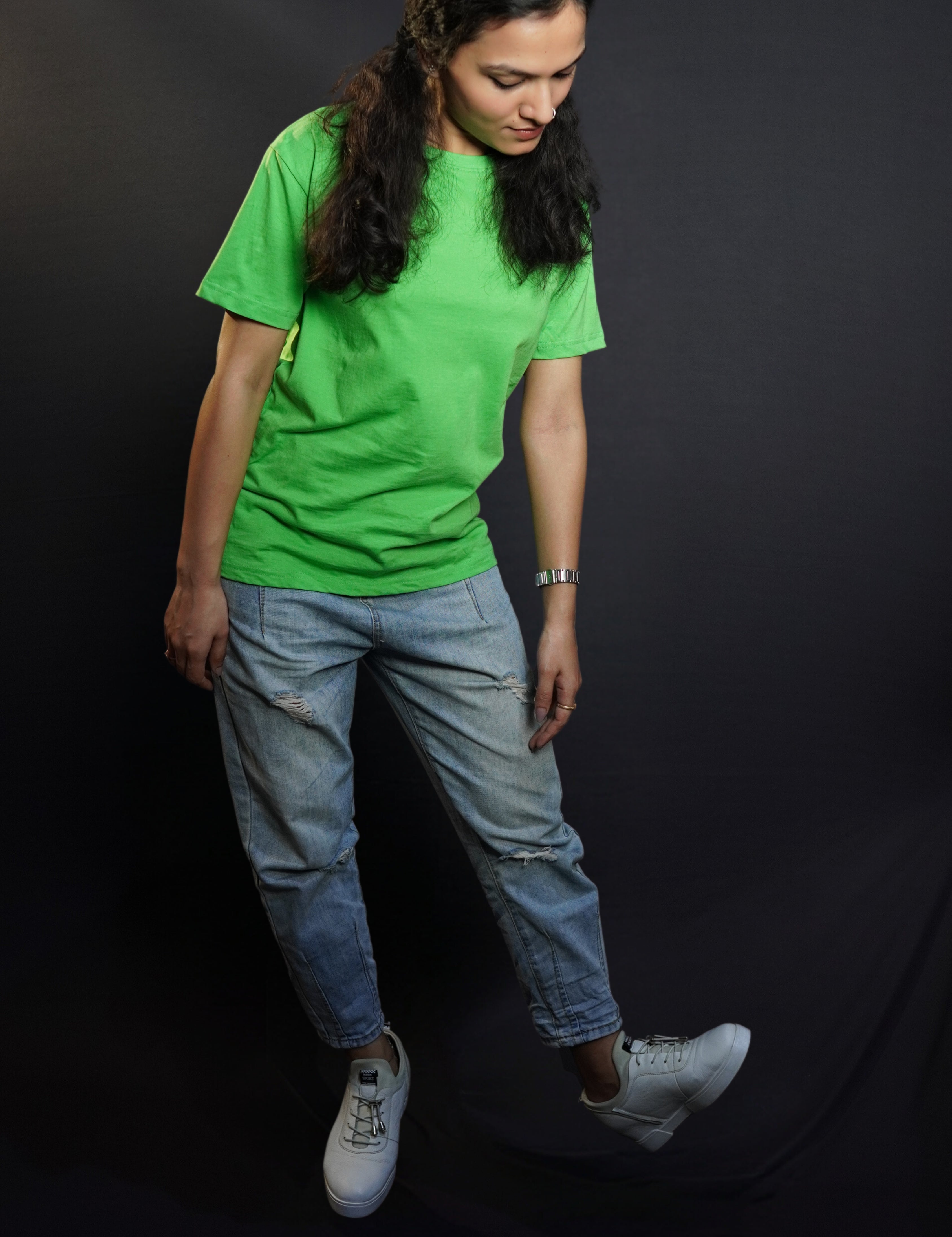 A woman wearing a women's long length short sleeved stretchable liril green t-shirt, posing against a dark background as she bends down slightly, looking at her white shoes, showcasing a relaxed cool fit-Muselot