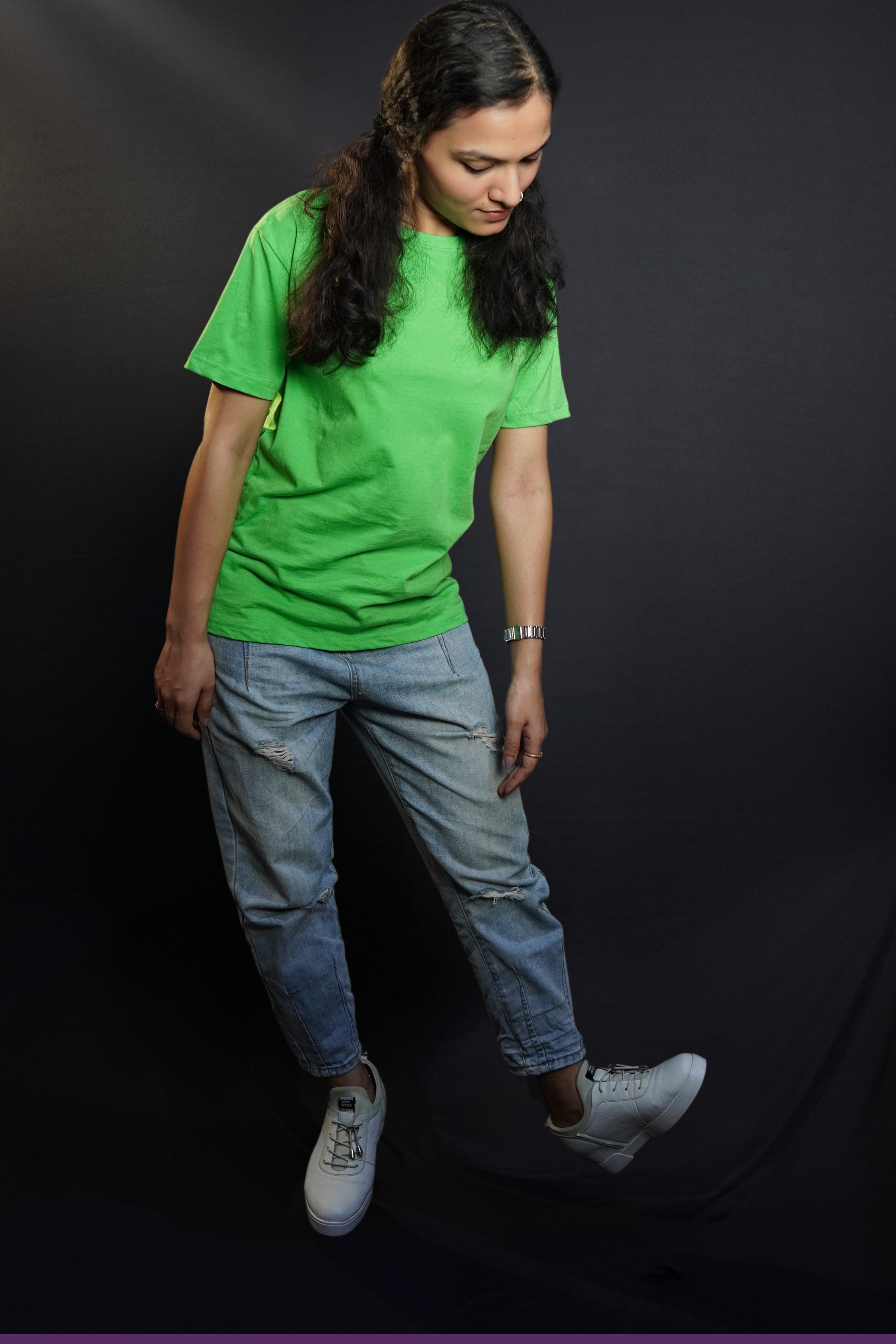A woman wearing a women's long length short sleeved stretchable liril green t-shirt, posing against a dark background as she bends down slightly, looking at her white shoes, showcasing a relaxed cool fit-Muselot