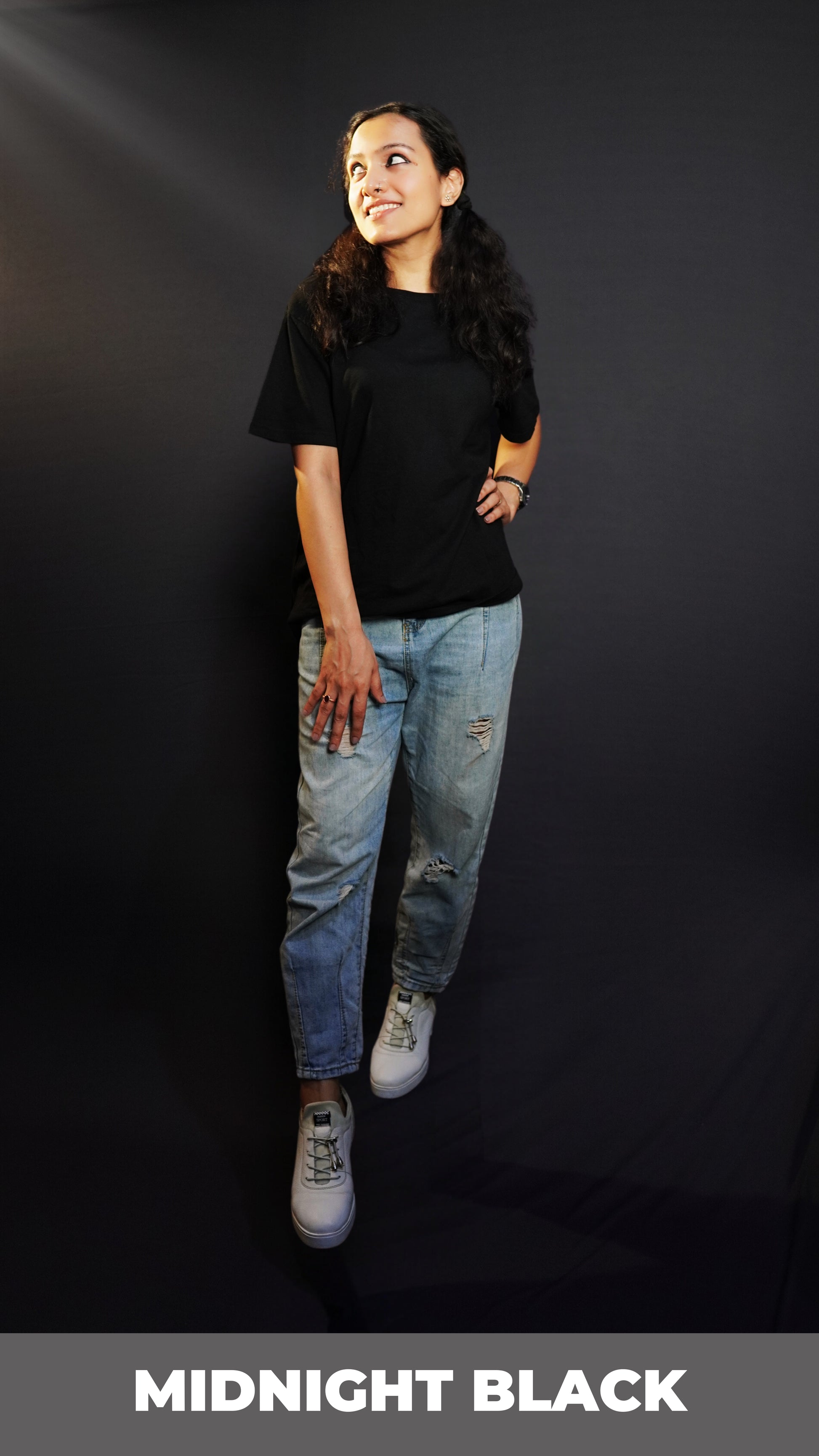 An elegant model wearing a short sleeved long length plain midnight black women's t-shirt, looking sideways as her hand gently lies on her waist and thigh, highlighting a stylish modern look-Muselot