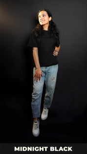 An elegant model wearing a short sleeved long length plain midnight black women's t-shirt, looking sideways as her hand gently lies on her waist and thigh, highlighting a stylish modern look-Muselot