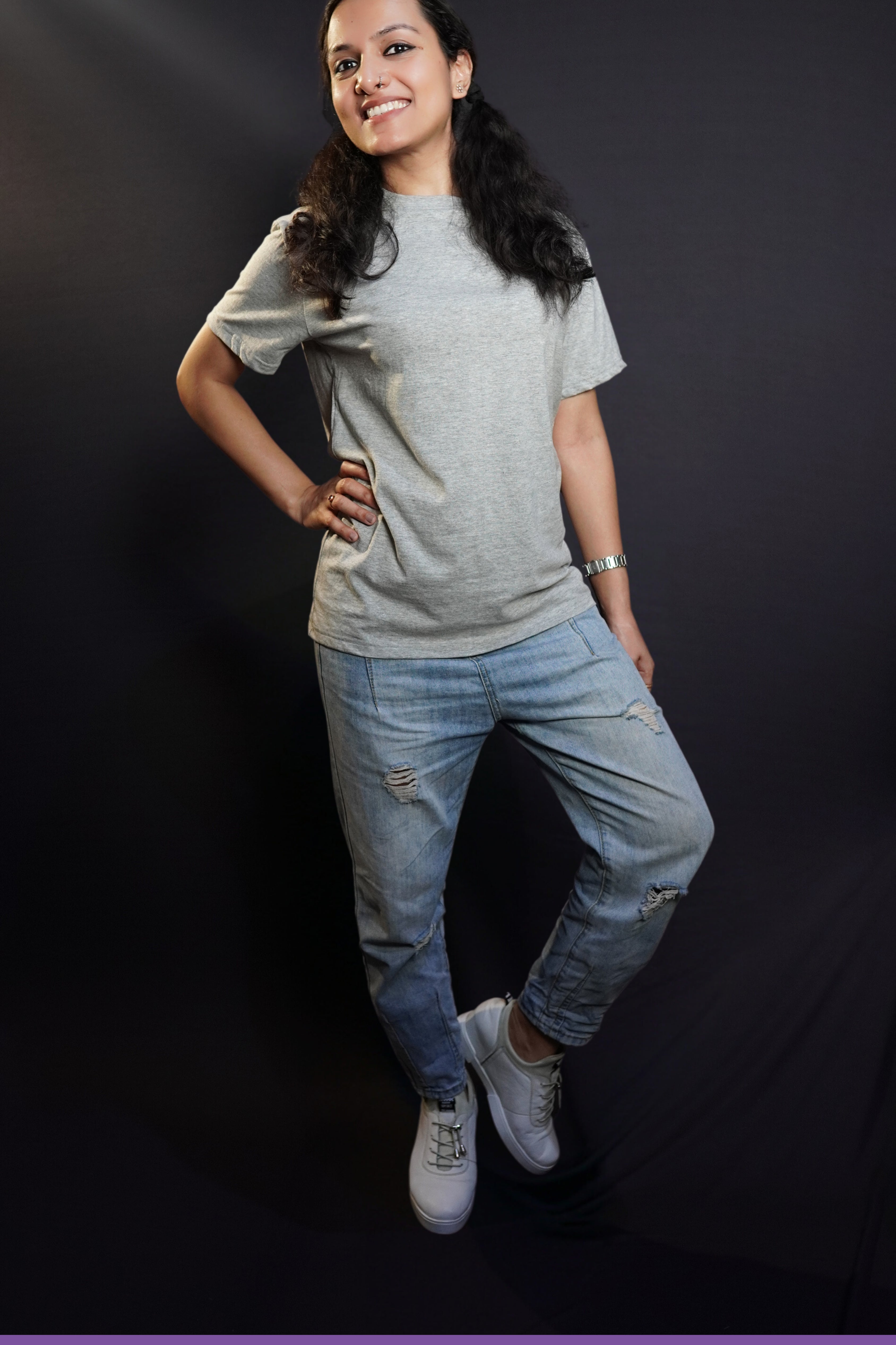 A happy woman posing with her hand on her waist and a leg bending on her side, wearing a men's plain long length misty grey t-shirt, showcasing a funky snug fit-Muselot