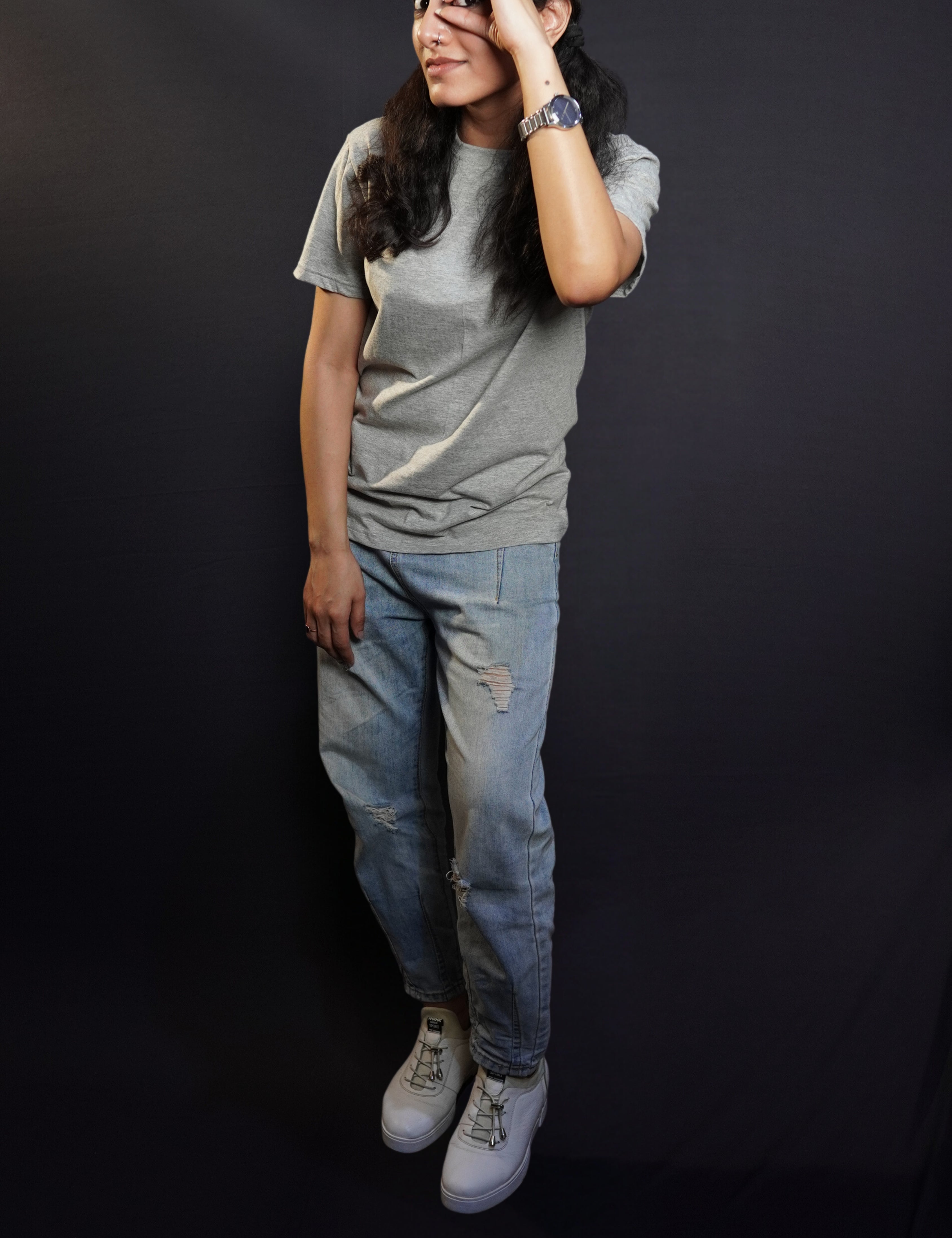 A woman wearing a plus size moisture wicking plain round neck misty grey t-shirt, posing with her hand making a circle with a finder and thumb upon her eye, standing against a dark background, showcasing a cool informal look-Muselot
