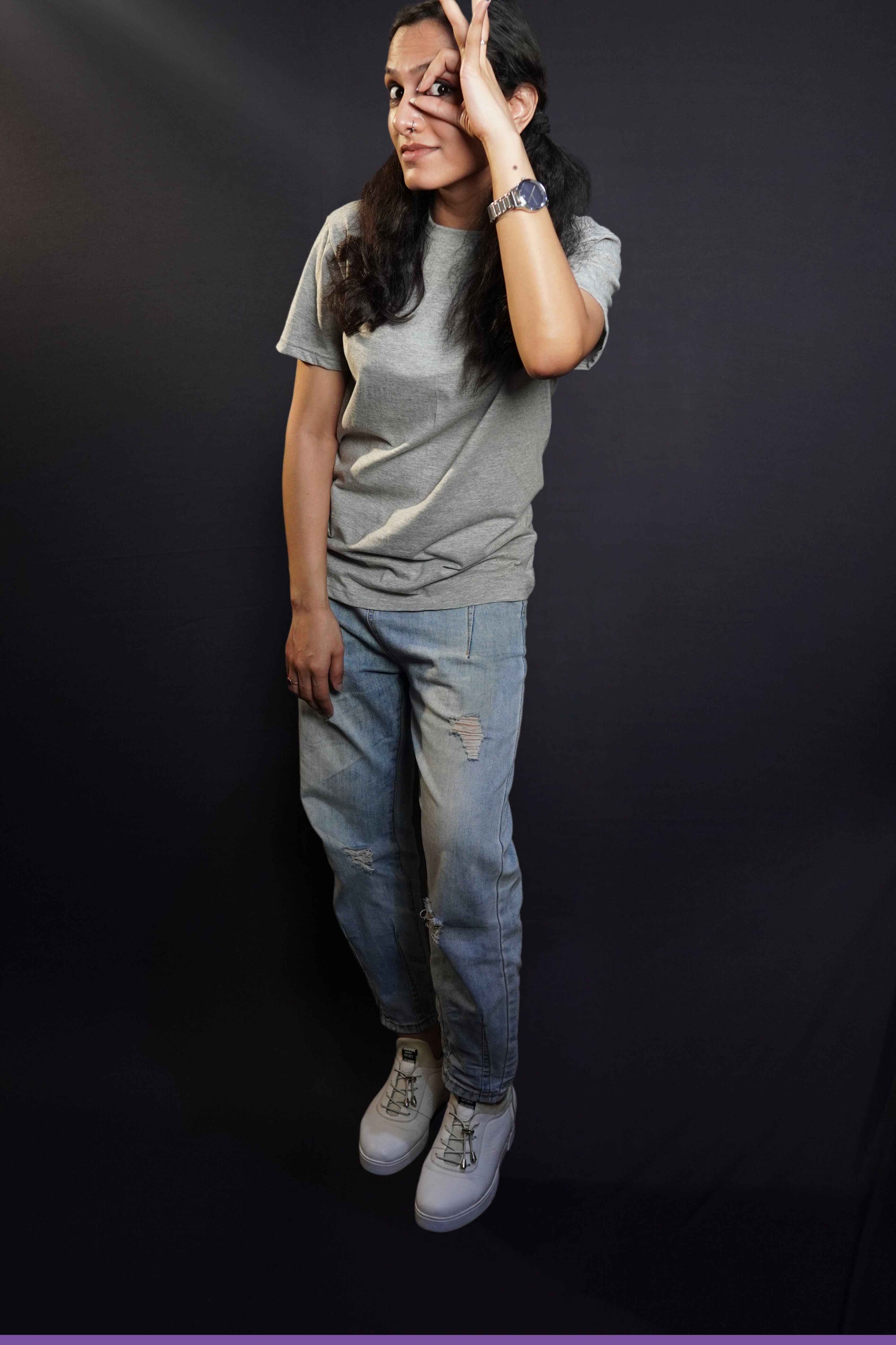A woman wearing a plus size moisture wicking plain round neck misty grey t-shirt, posing with her hand making a circle with a finder and thumb upon her eye, standing against a dark background, showcasing a cool informal look-Muselot