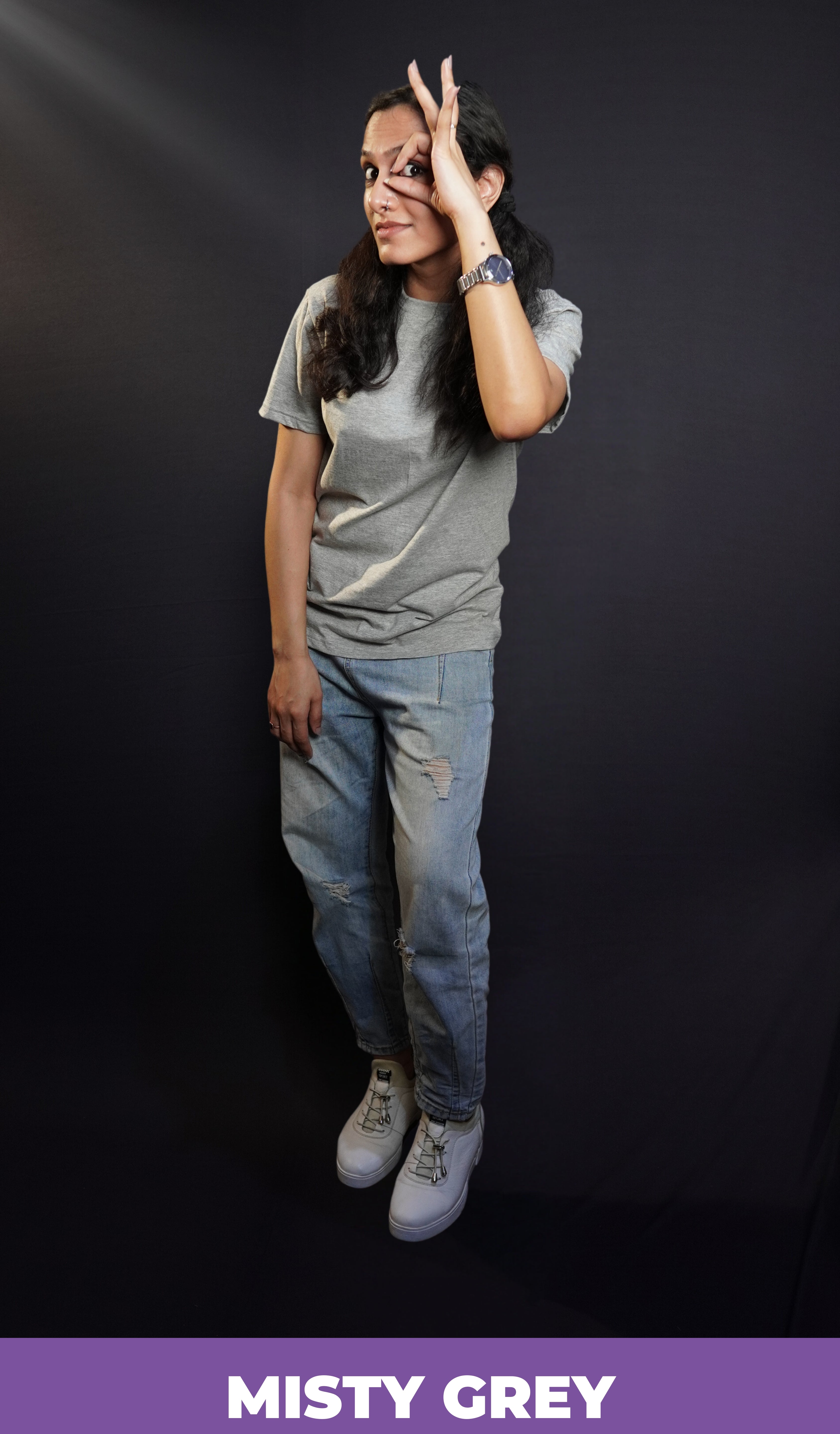 A woman wearing a plus size moisture wicking plain round neck misty grey t-shirt, posing with her hand making a circle with a finder and thumb upon her eye, standing against a dark background, showcasing a cool informal look-Muselot