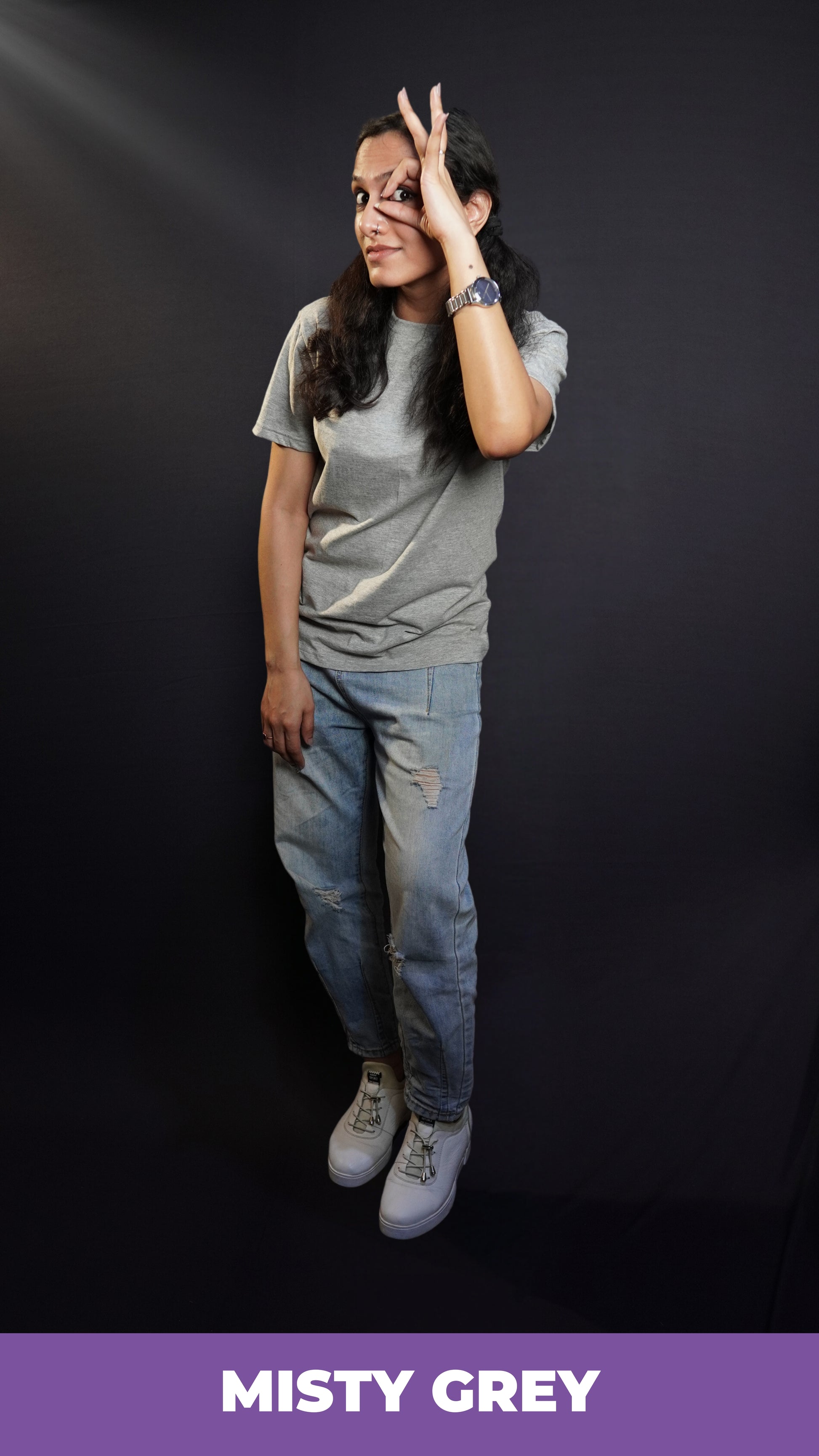 A woman wearing a plus size moisture wicking plain round neck misty grey t-shirt, posing with her hand making a circle with a finder and thumb upon her eye, standing against a dark background, showcasing a cool informal look-Muselot