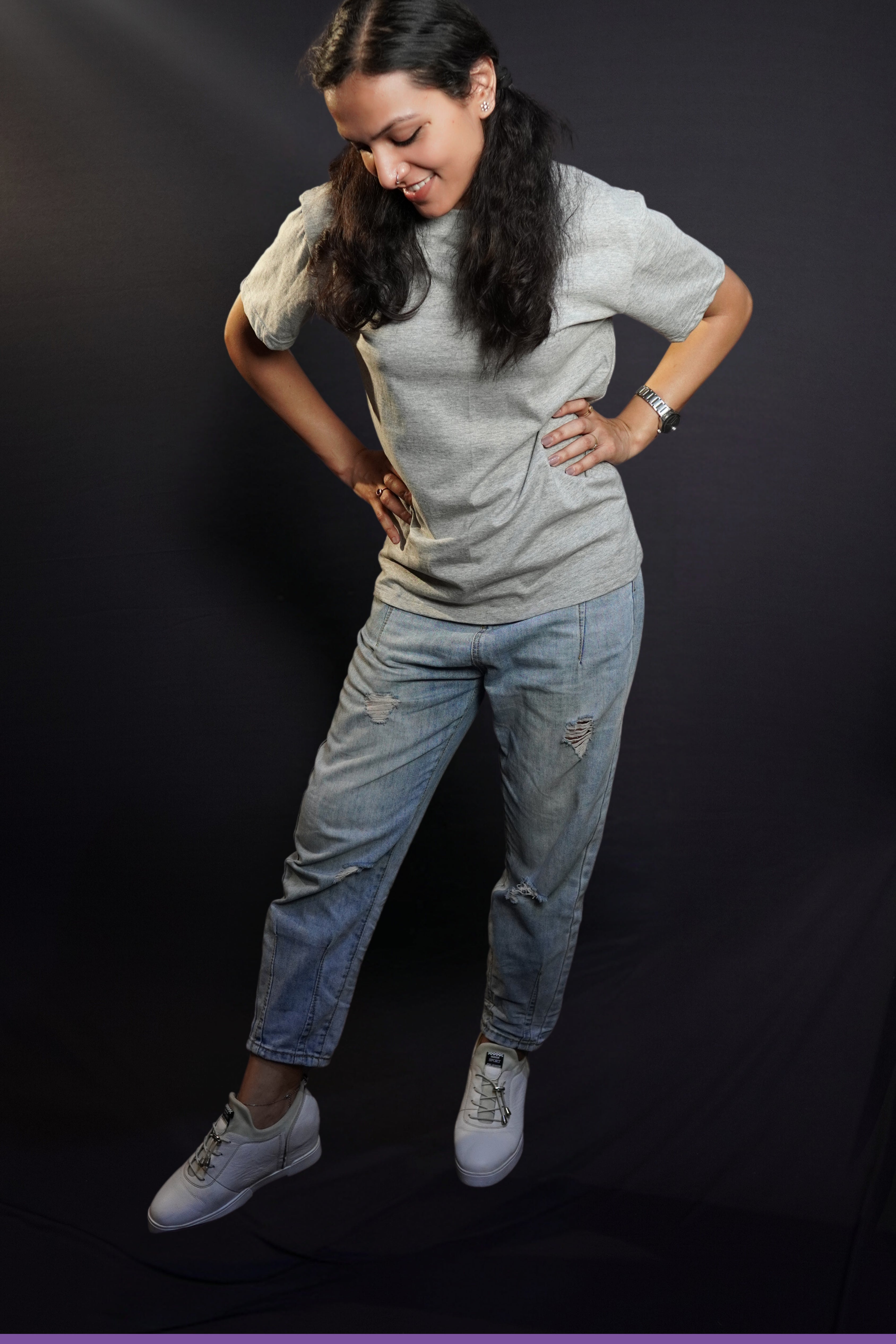 A woman posing with her hands on her waist standing slghtly sideways bending down a little, wearing a plain unisex short sleeved stretchable misty grey t-shirt, highlighting a stylish comfortable fit-Muselot