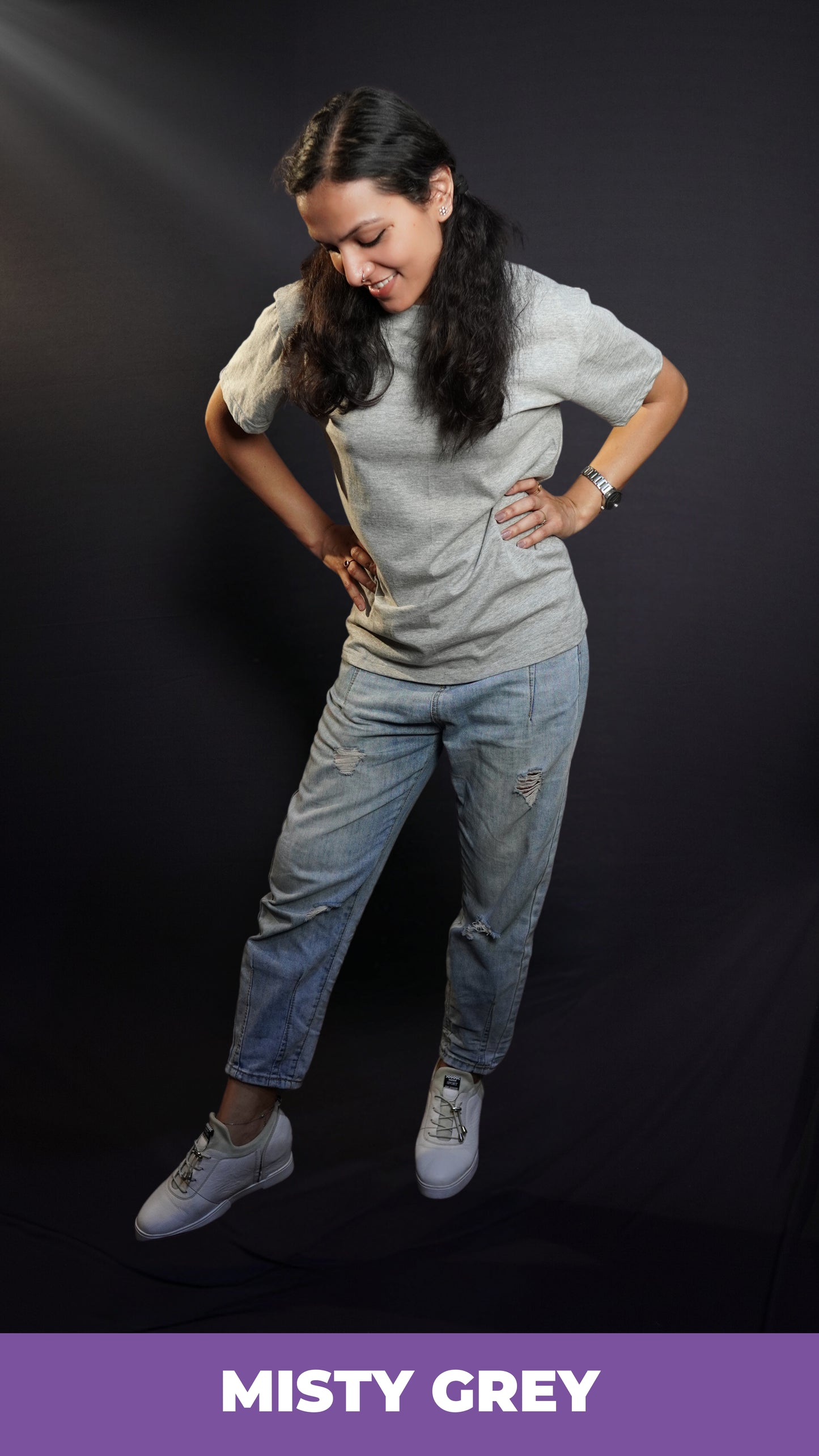 A woman posing with her hands on her waist standing slghtly sideways bending down a little, wearing a plain unisex short sleeved stretchable misty grey t-shirt, highlighting a stylish comfortable fit-Muselot