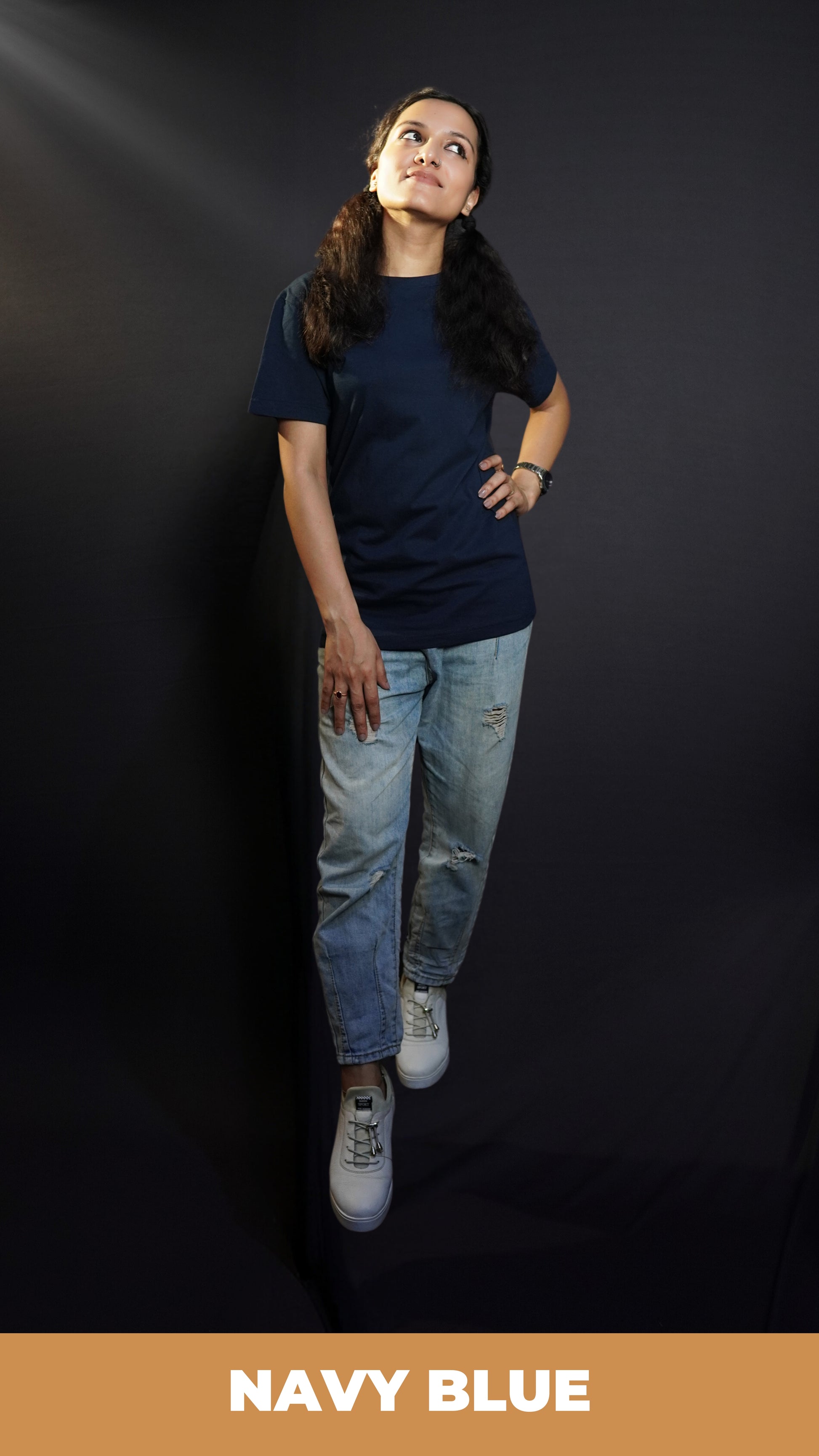 An elegant model wearing a women's cotton round neck stretchable plain navy blue t-shirt, posing with her hand on her waist and other on her thigh as she looks sideways with her chin up high, showcasing a popular modish look-Muselot