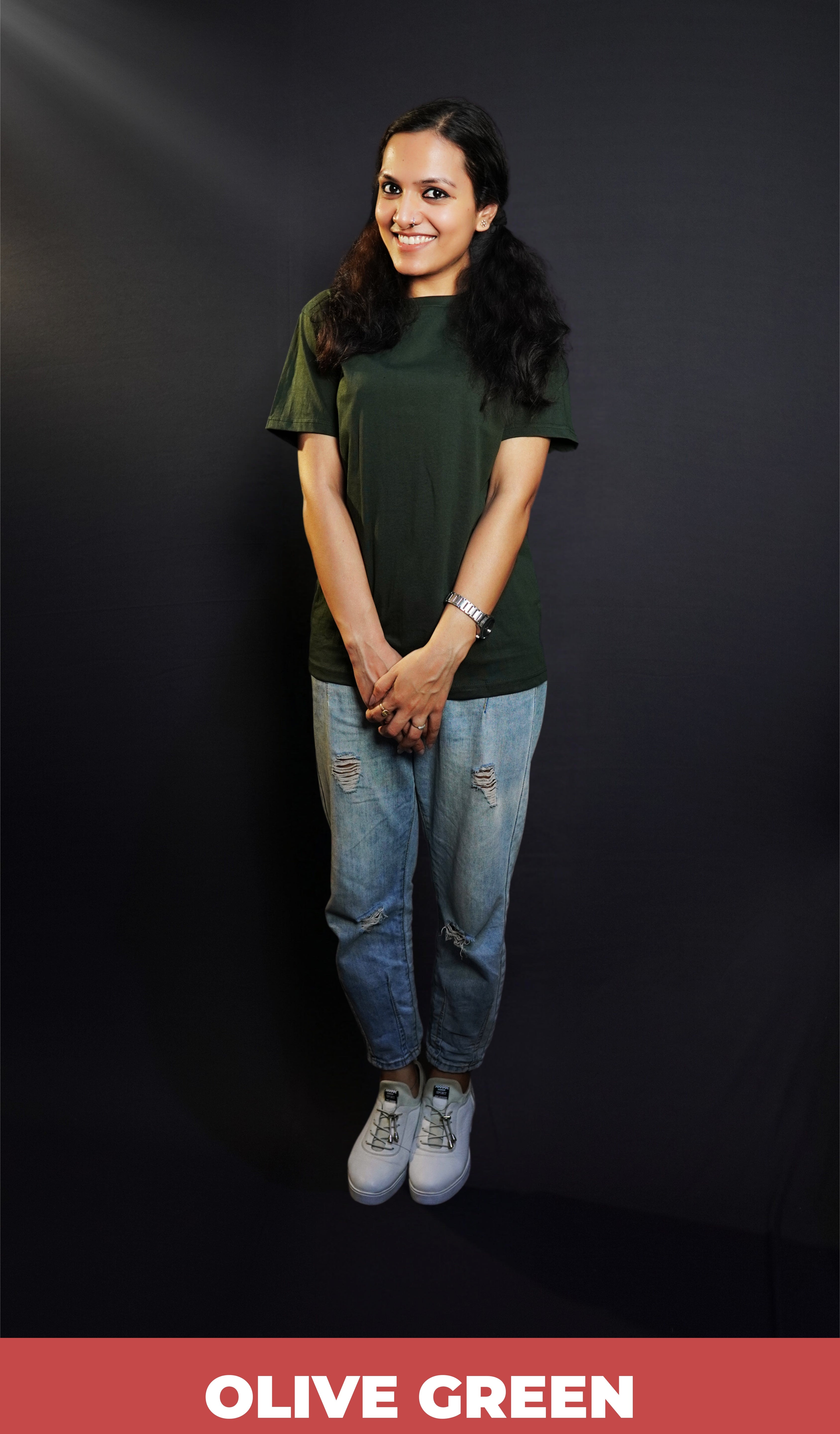 A woman happily smiling standing against a dark background with her hand grabbing the other, wearing a moisture wicking stretchable plain cotton olive green t-shirt, highlighting a flowing aesthetic look-Muselot