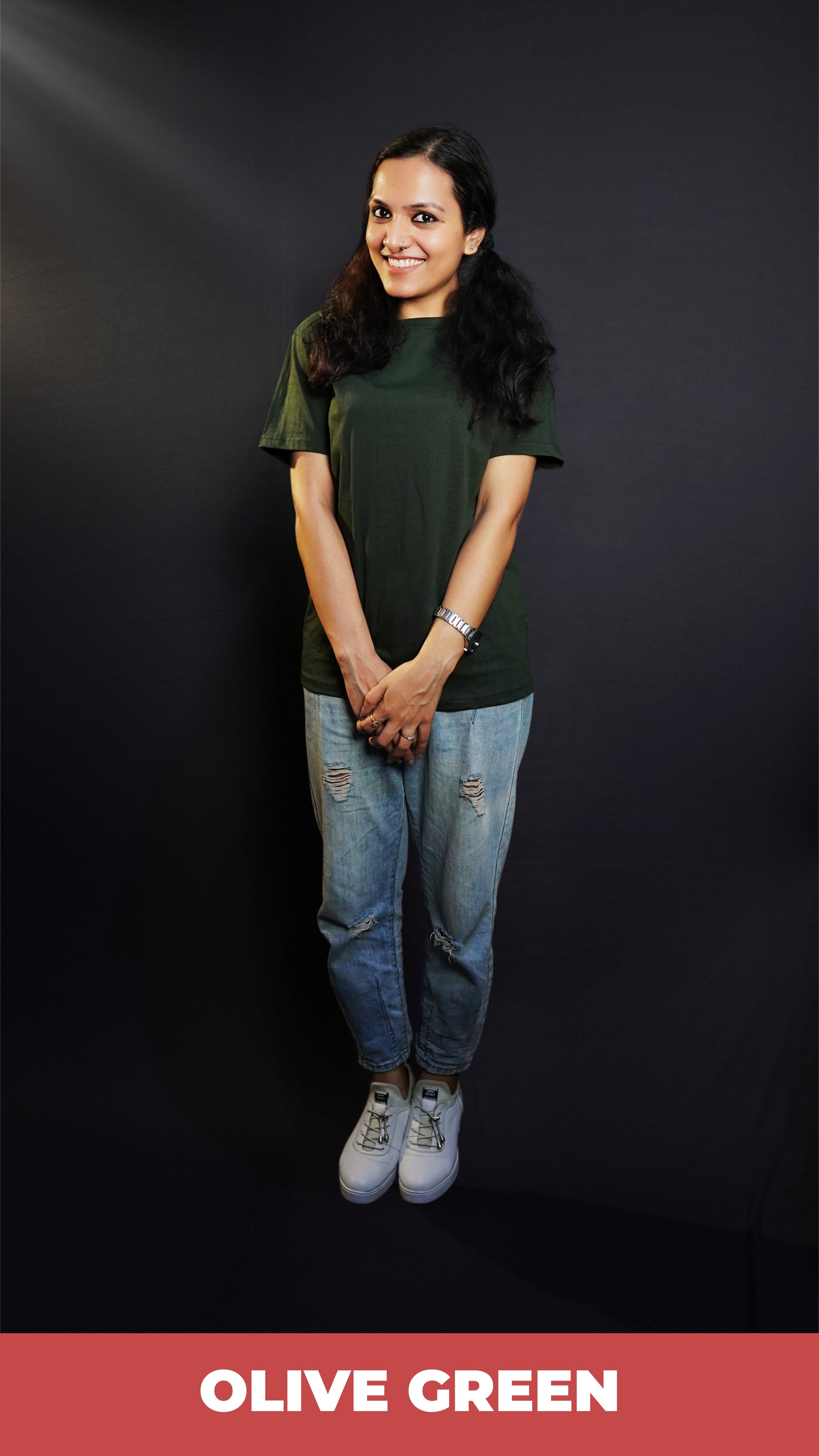 A woman happily smiling standing against a dark background with her hand grabbing the other, wearing a moisture wicking stretchable plain cotton olive green t-shirt, highlighting a flowing aesthetic look-Muselot