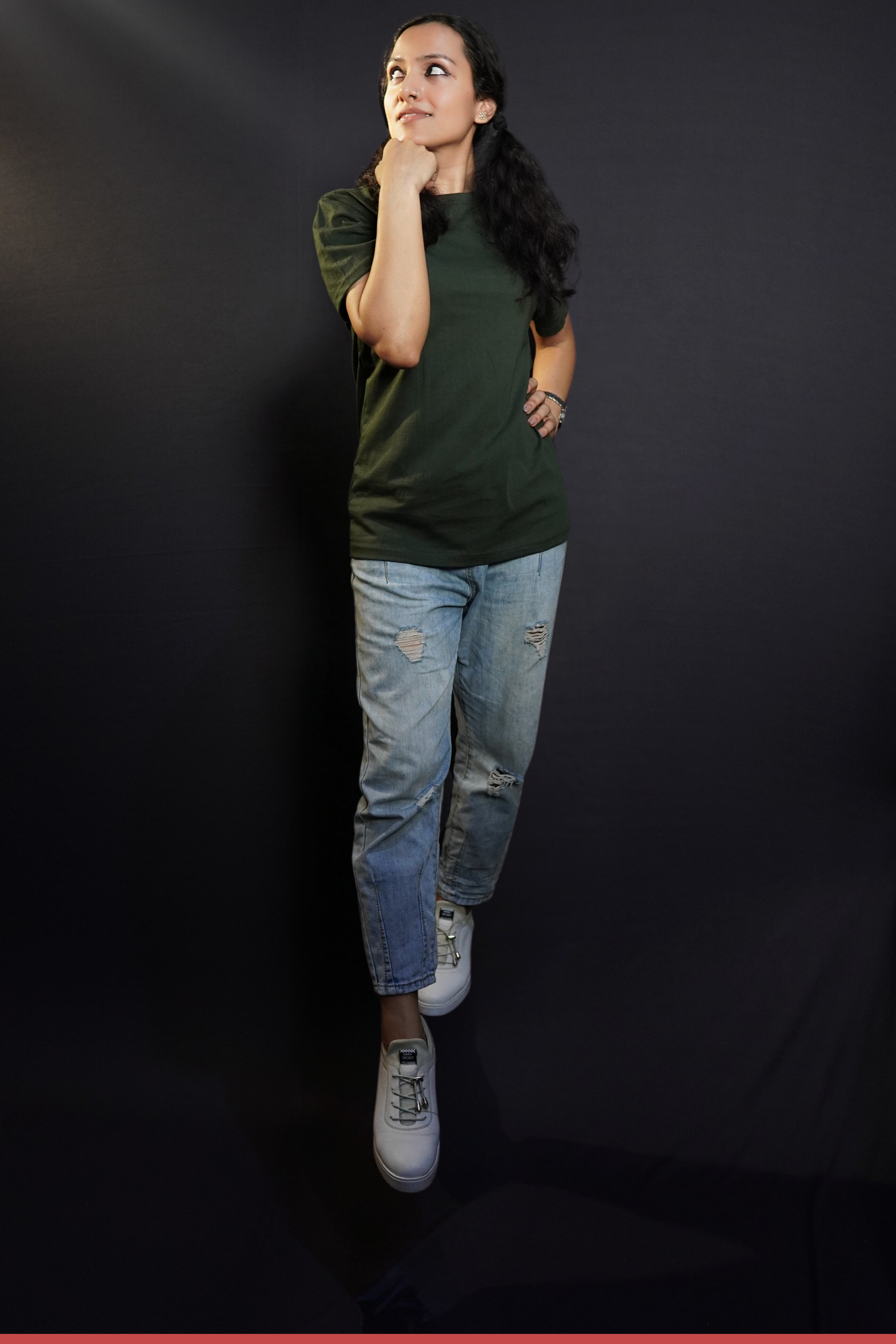 A model wearing a round neck stretchable plain olive green t-shirt, posing with her fist under her chin and other on her waist as she glances sideways with a small smile on her lips, highlighting a laid back everyday look-Muselot