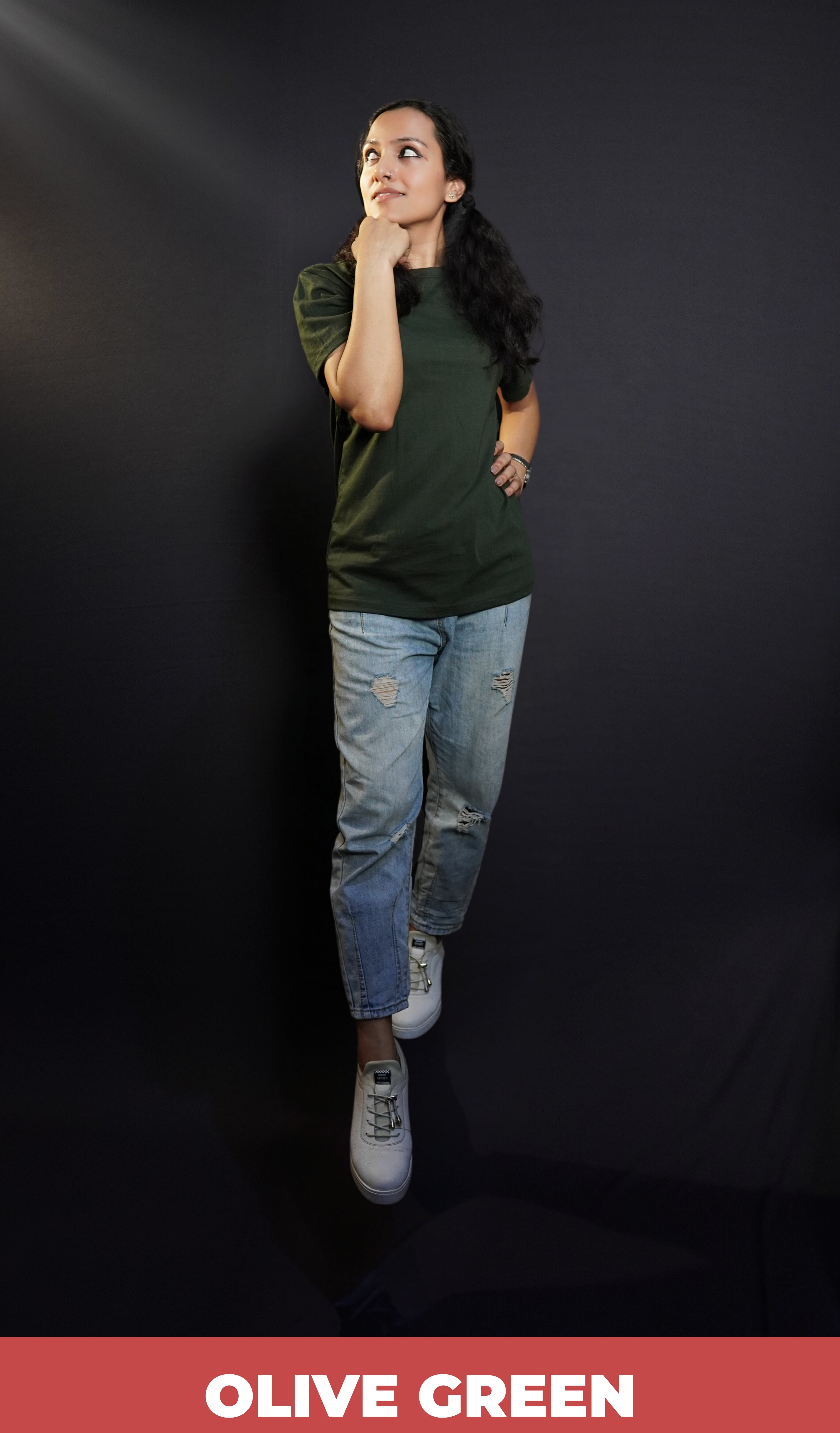 A model wearing a round neck stretchable plain olive green t-shirt, posing with her fist under her chin and other on her waist as she glances sideways with a small smile on her lips, highlighting a laid back everyday look-Muselot