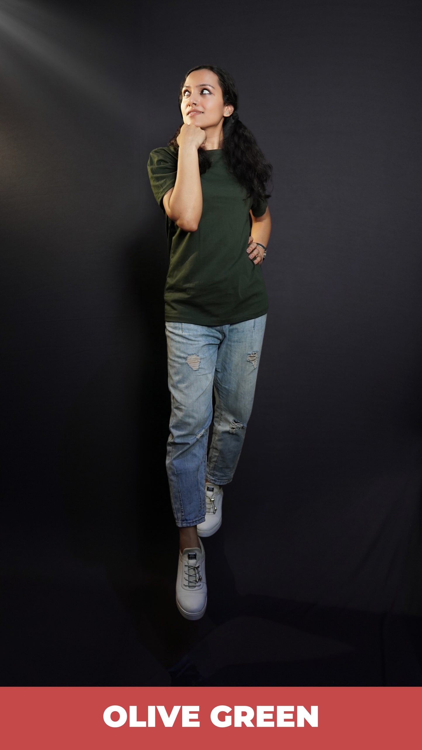 A model wearing a round neck stretchable plain olive green t-shirt, posing with her fist under her chin and other on her waist as she glances sideways with a small smile on her lips, highlighting a laid back everyday look-Muselot