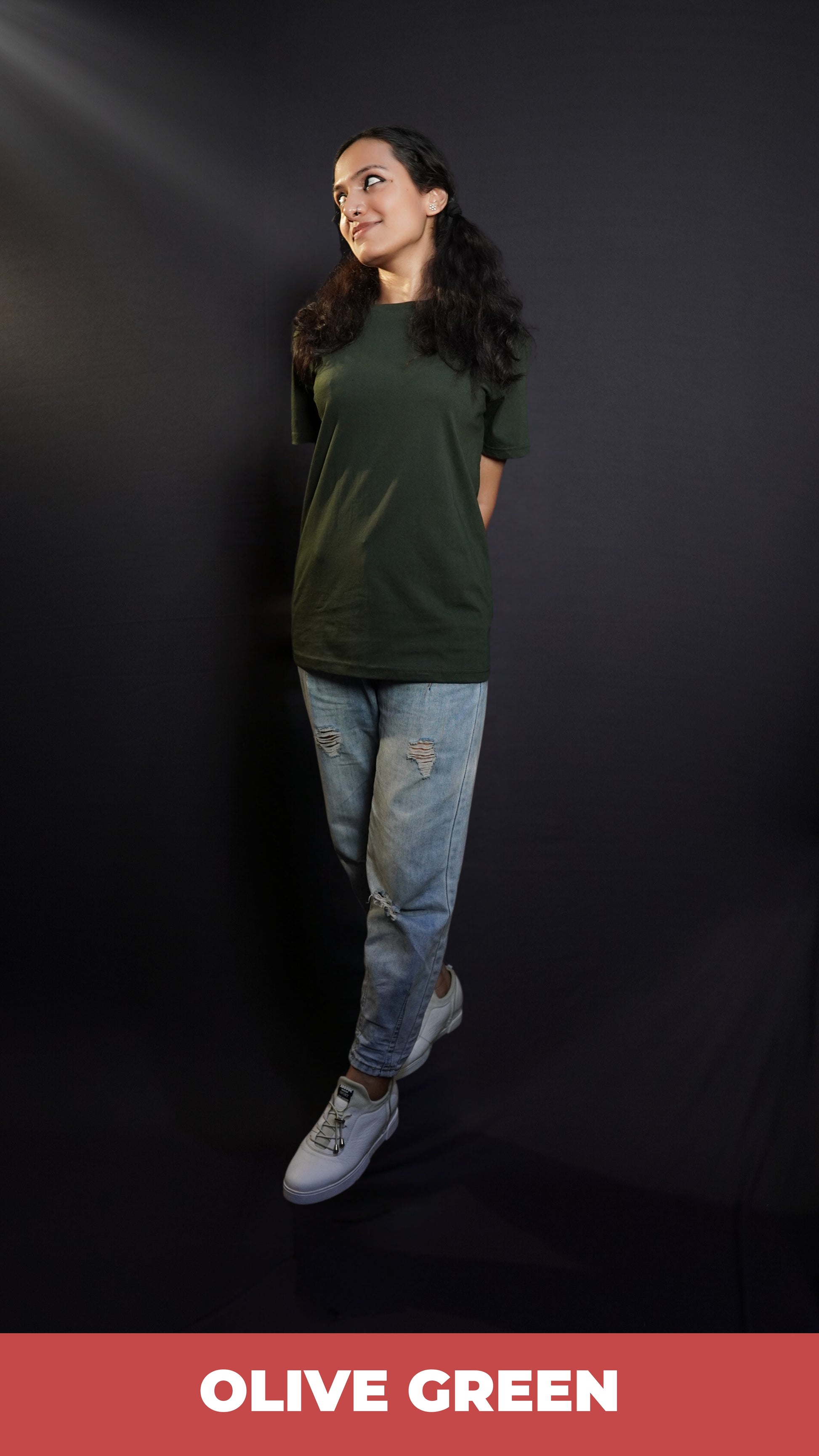 A stylish model posing with her hands at her back as she walks against a dark background, wearing a unisex plain round neck olive green t-shirt, showcasing a trendy snug fit-Muselot