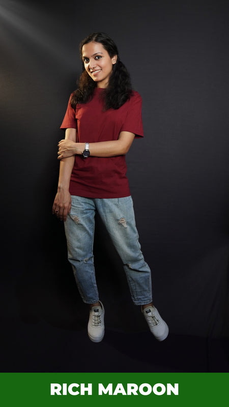 A woman wearing a plus size short sleeved moisture wicking rich maroon t-shirt, posing with her hand grabbing her elbow, smiling happily, highlighting a popular baggy fit-Muselot