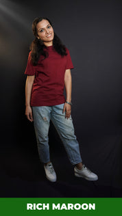 A woman wearing a long length summer cotton rich maroon t-shirt, posing with her hand lying gently on her thigh as she stands against a dark background, showcasing a modish easygoing fit-Muselot