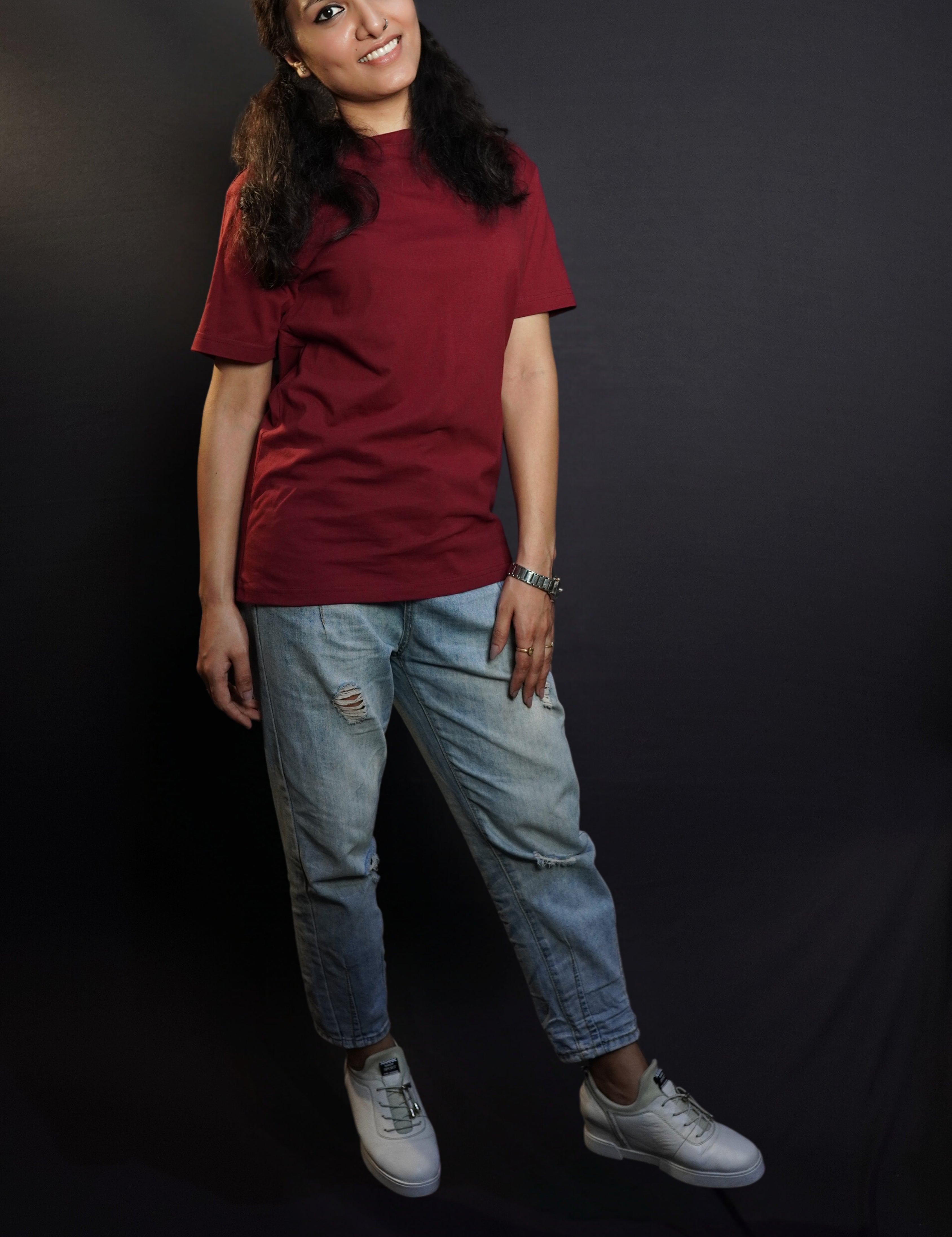 A woman wearing a long length summer cotton rich maroon t-shirt, posing with her hand lying gently on her thigh as she stands against a dark background, showcasing a modish easygoing fit-Muselot