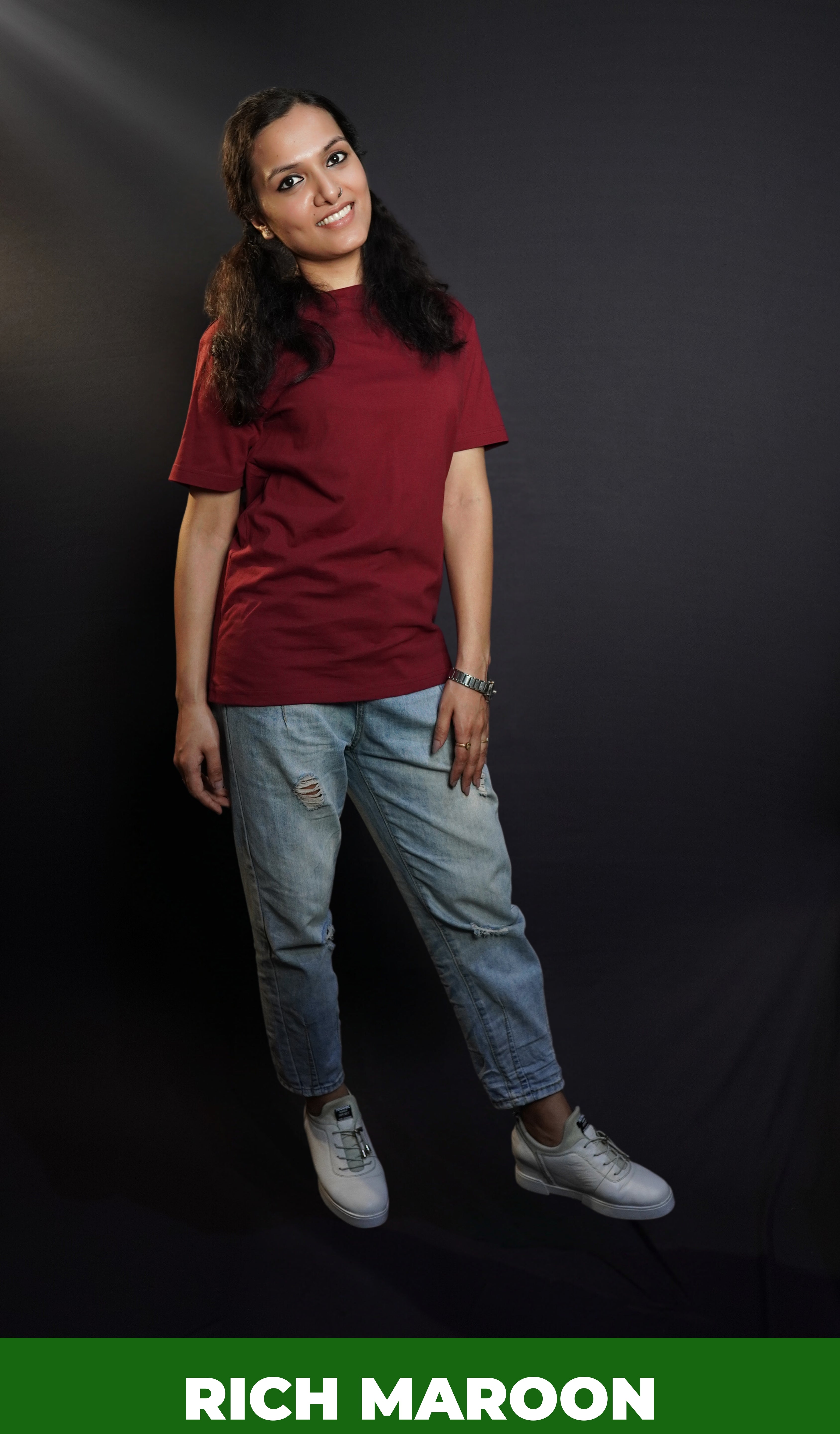 A woman wearing a long length summer cotton rich maroon t-shirt, posing with her hand lying gently on her thigh as she stands against a dark background, showcasing a modish easygoing fit-Muselot