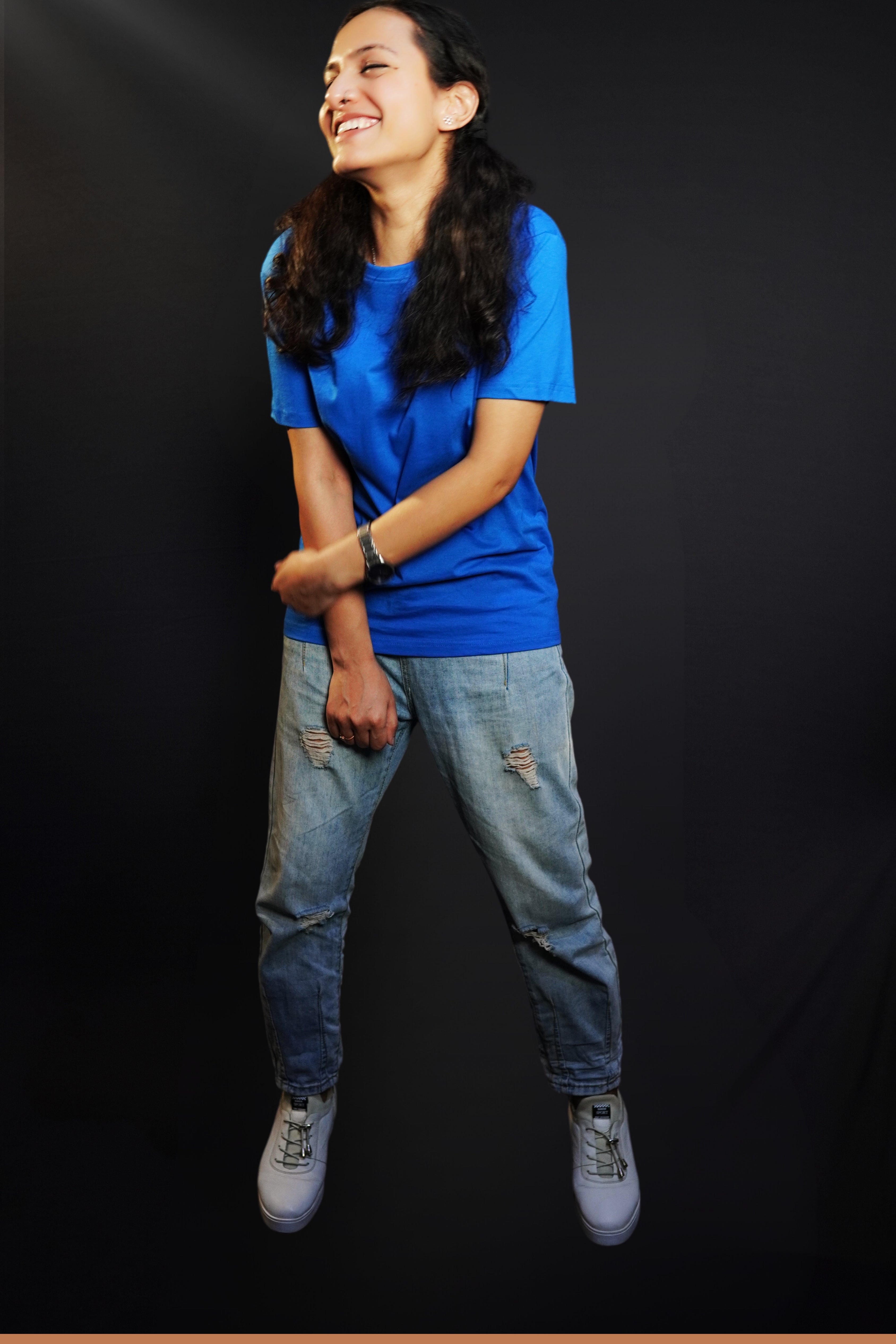 A woman laughing out loud with her hand grabbing her forearm standing against a dark background, wearing a long length cotton quick drying royal blue t-shirt, highlighting an in demand relaxed look-Muselot