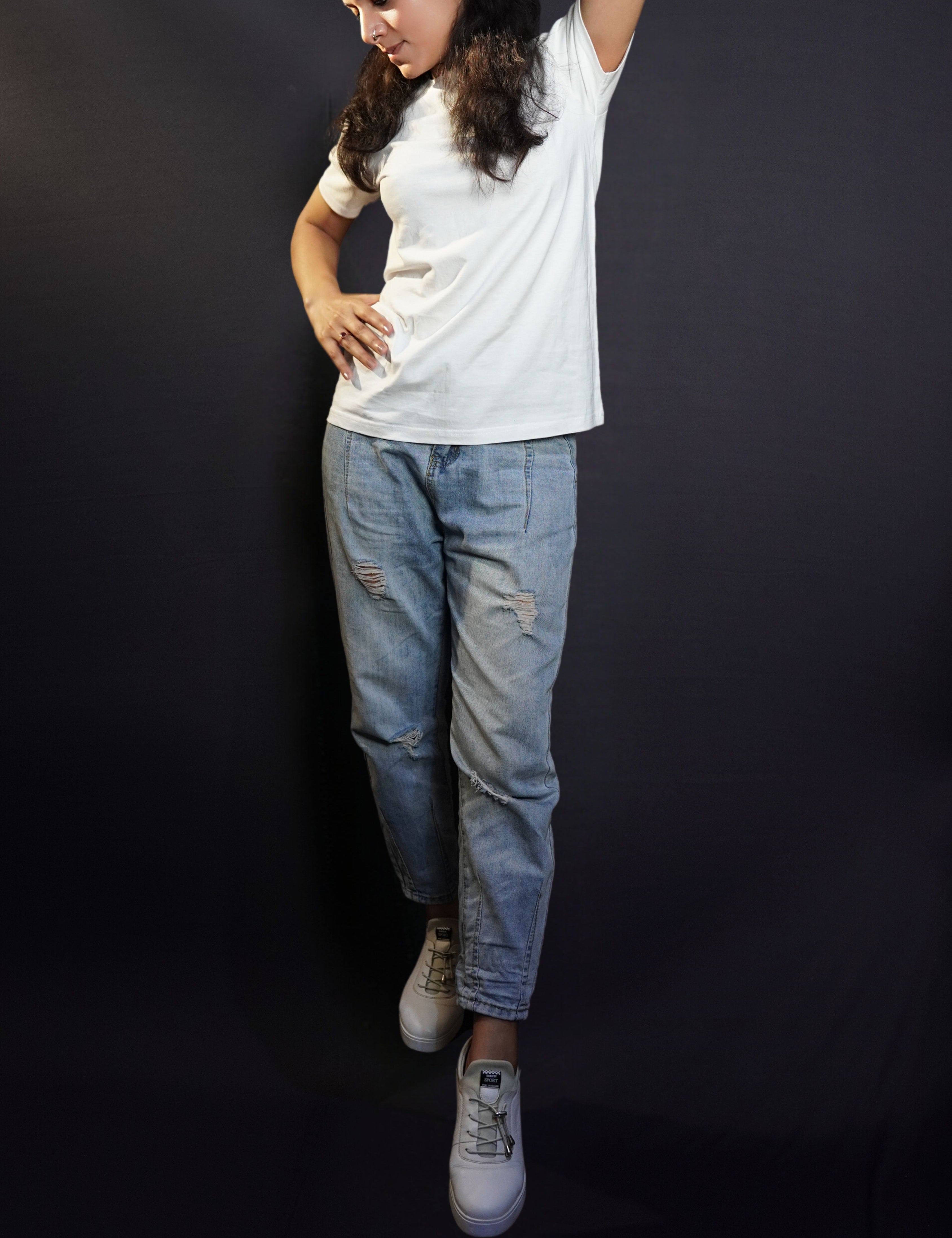 A woman posing with her hand up behind her neck and another on her waist as she looks down at the dark ground, wearing a short sleeved moisture wicking stretchable cotton snowy white t-shirt, showcasing a stylish in vogue look-Muselot