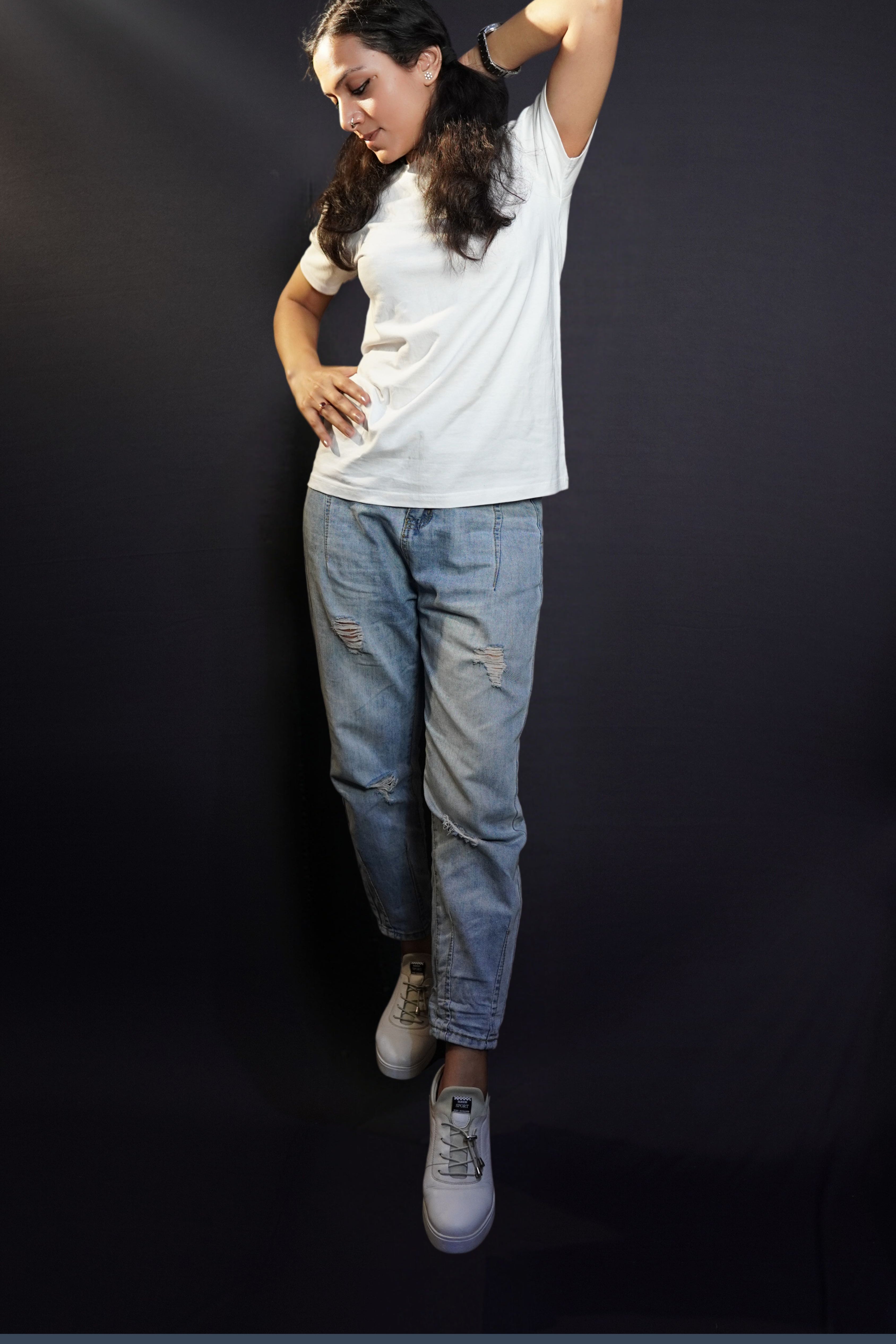 A woman posing with her hand up behind her neck and another on her waist as she looks down at the dark ground, wearing a short sleeved moisture wicking stretchable cotton snowy white t-shirt, showcasing a stylish in vogue look-Muselot