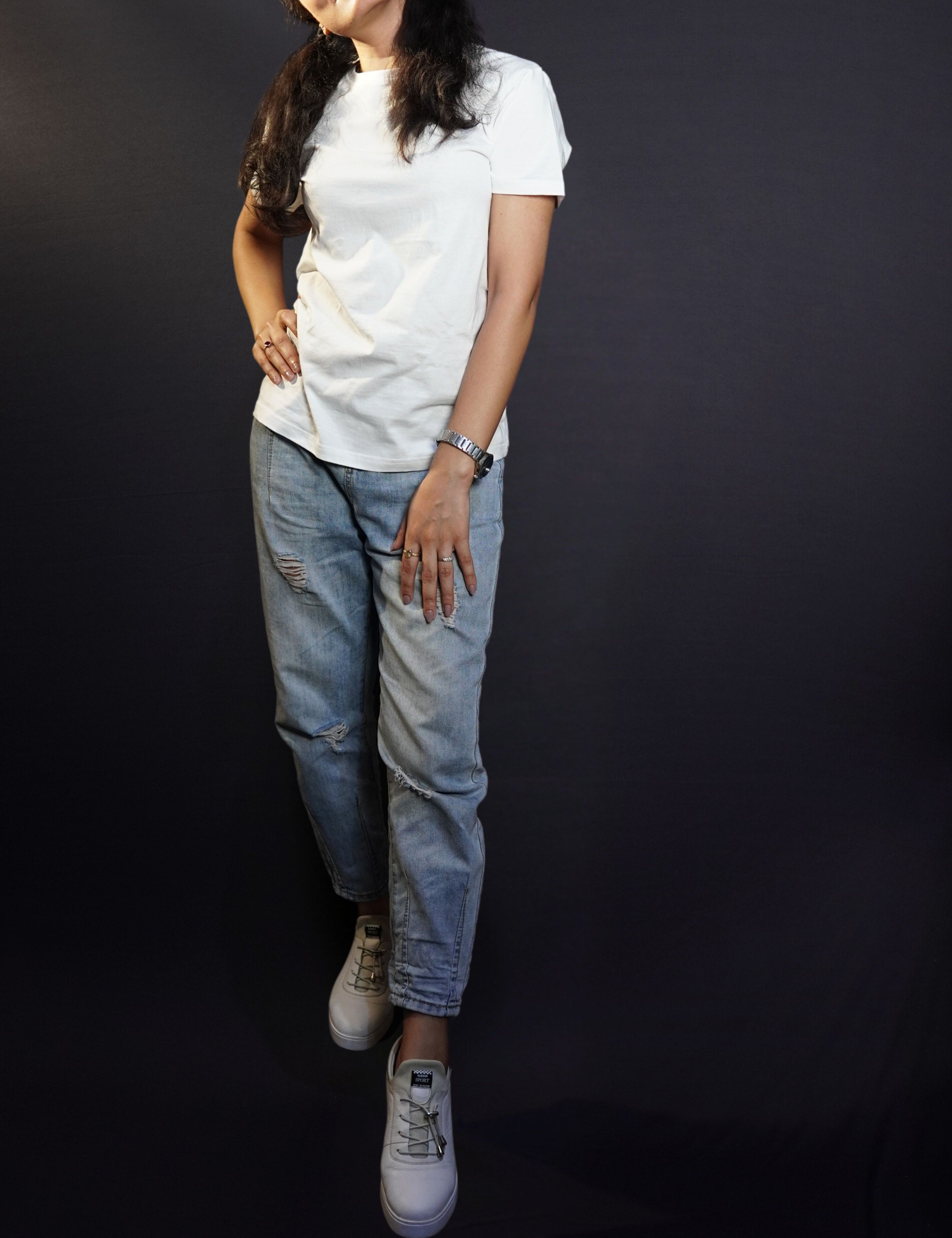 A woman looking sideways with her hand on her thigh and another on her waist standing slightly sideways against a dark backgroud, wearing a snowy white plain short sleeved round neck t-shirt, showcasing a cool summer look-Muselot