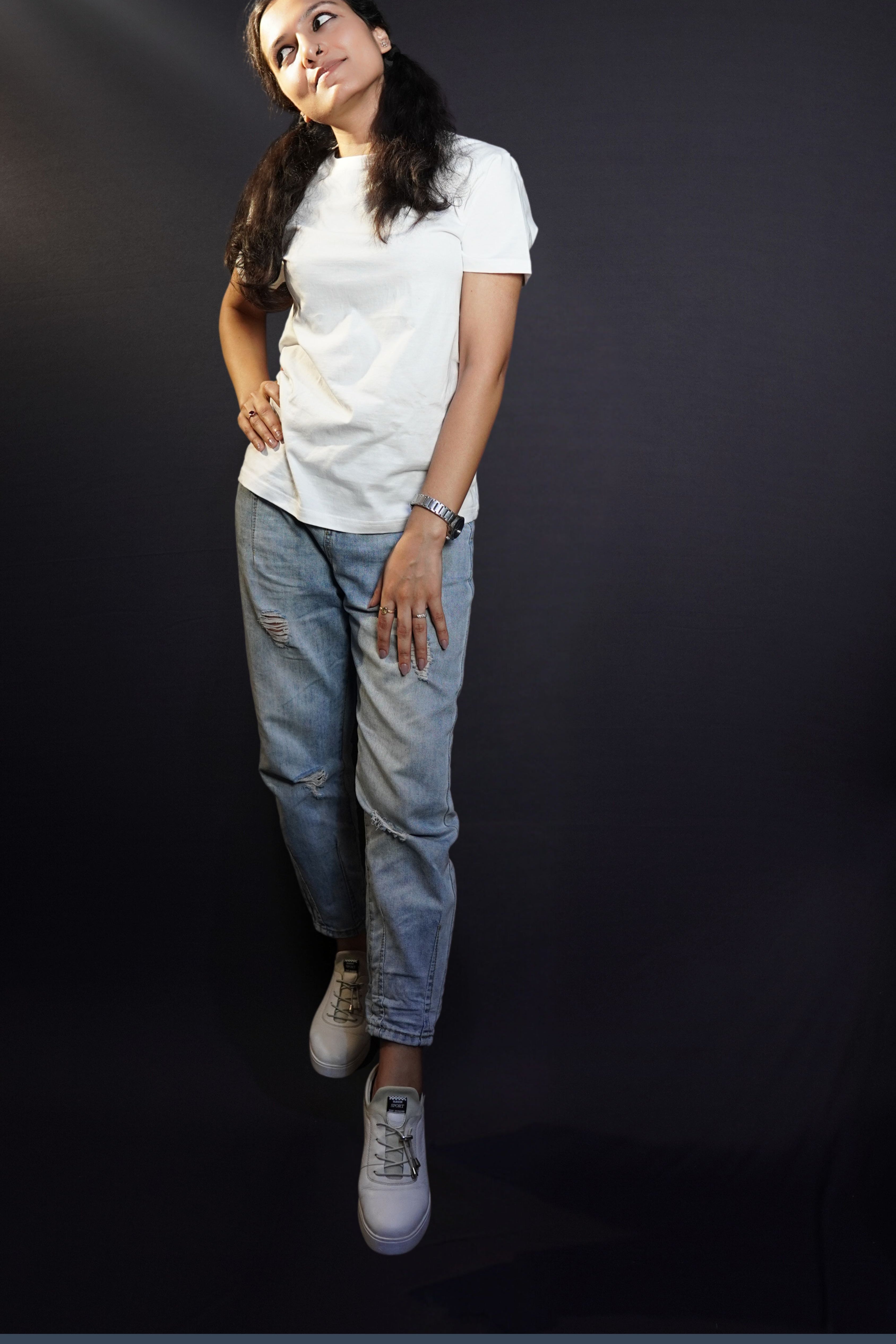 A woman looking sideways with her hand on her thigh and another on her waist standing slightly sideways against a dark backgroud, wearing a snowy white plain short sleeved round neck t-shirt, showcasing a cool summer look-Muselot
