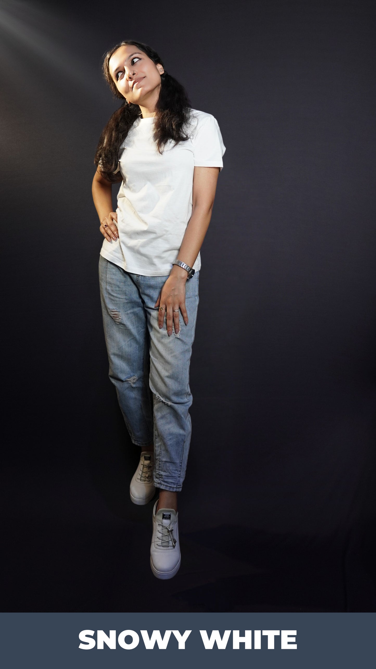 A woman looking sideways with her hand on her thigh and another on her waist standing slightly sideways against a dark backgroud, wearing a snowy white plain short sleeved round neck t-shirt, showcasing a cool summer look-Muselot