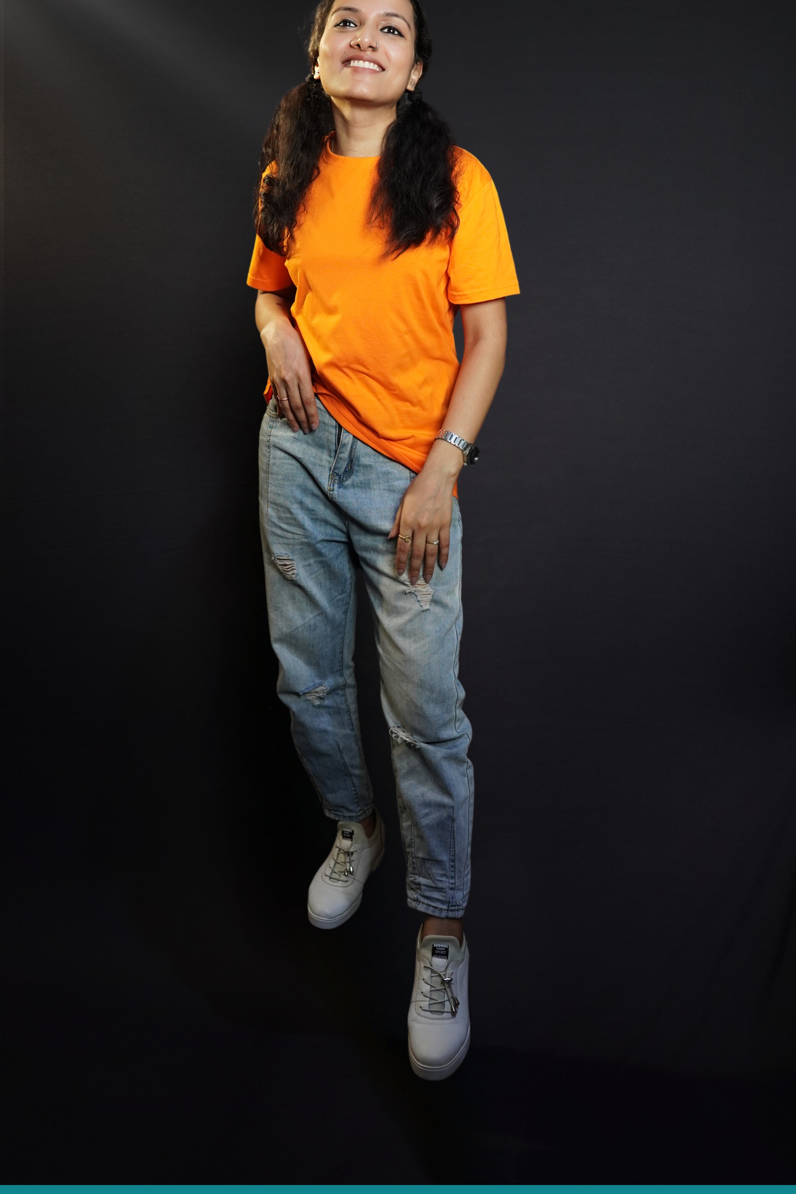 A stylish model posing happily as her hand is slightly inside her front pocket of her jeans, smiling happily, wearing a summer cotton plain tropical orange long length t-shirt, highlighting a cool modern look-Muselot